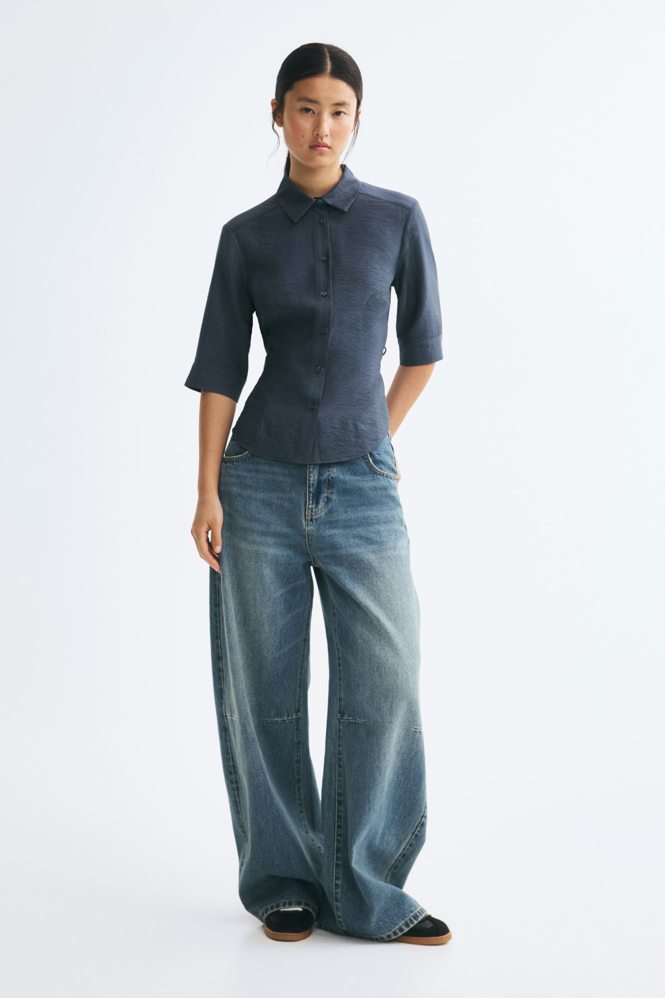 View larger image: A model is wearing a fitted, dark blue viscose-blend shirt, facing front, featuring a collar, a button-down front, and 3/4-length sleeves. The shirt creates a nipped-in silhouette at the waist and is styled with wide-leg denim jeans.