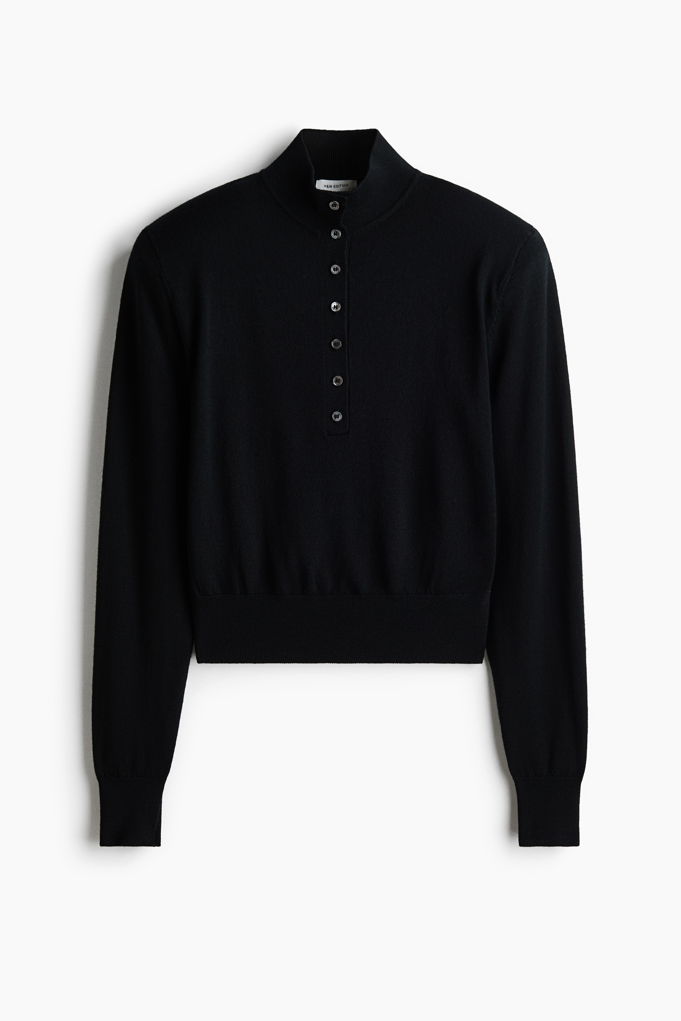 Un maglione nero in lana merino a maglia fine, caratterizzato da collo alto a coste, abbottonatura frontale e spalle strutturate, indossato dalla modella. La modella Ã¨ in piedi con le braccia incrociate, mettendo in risalto le maniche lunghe e i polsini a coste.