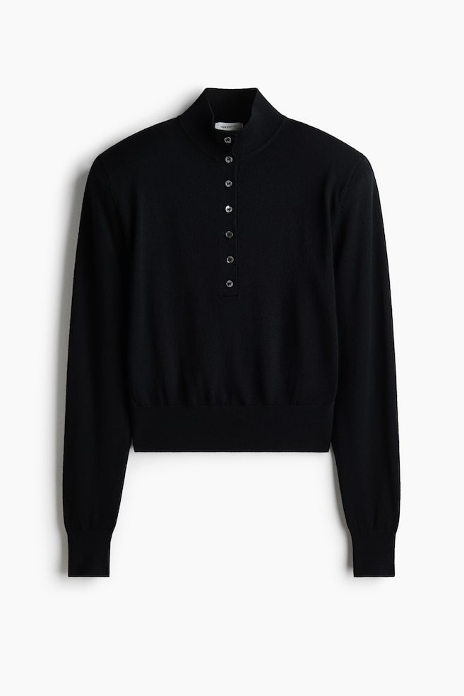 A black fine-knit sweater is shown from the front, featuring structured shoulders with shoulder pads, a ribbed high neck, and a central button placket. The sweater is displaying long sleeves with wide ribbed cuffs and a matching wide ribbed hem. - 1