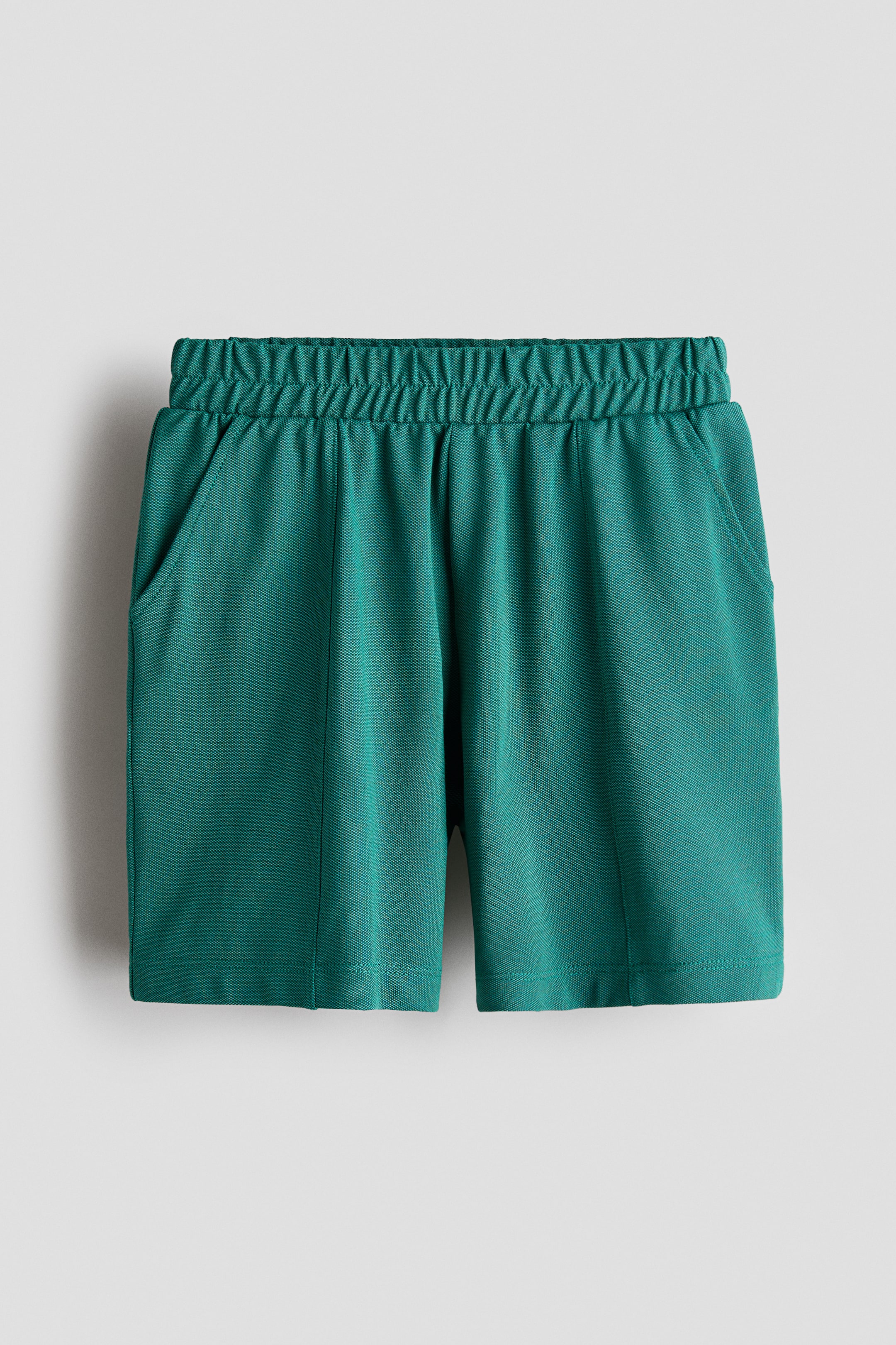 Xem ảnh lớn hơn: Chiếc quần short màu xanh ngọc lam này được chụp từ phía trước, thể hiện cạp chun nhún, túi xéo hai bên và các đường may ly dọc chạy xuống giữa mỗi ống quần.