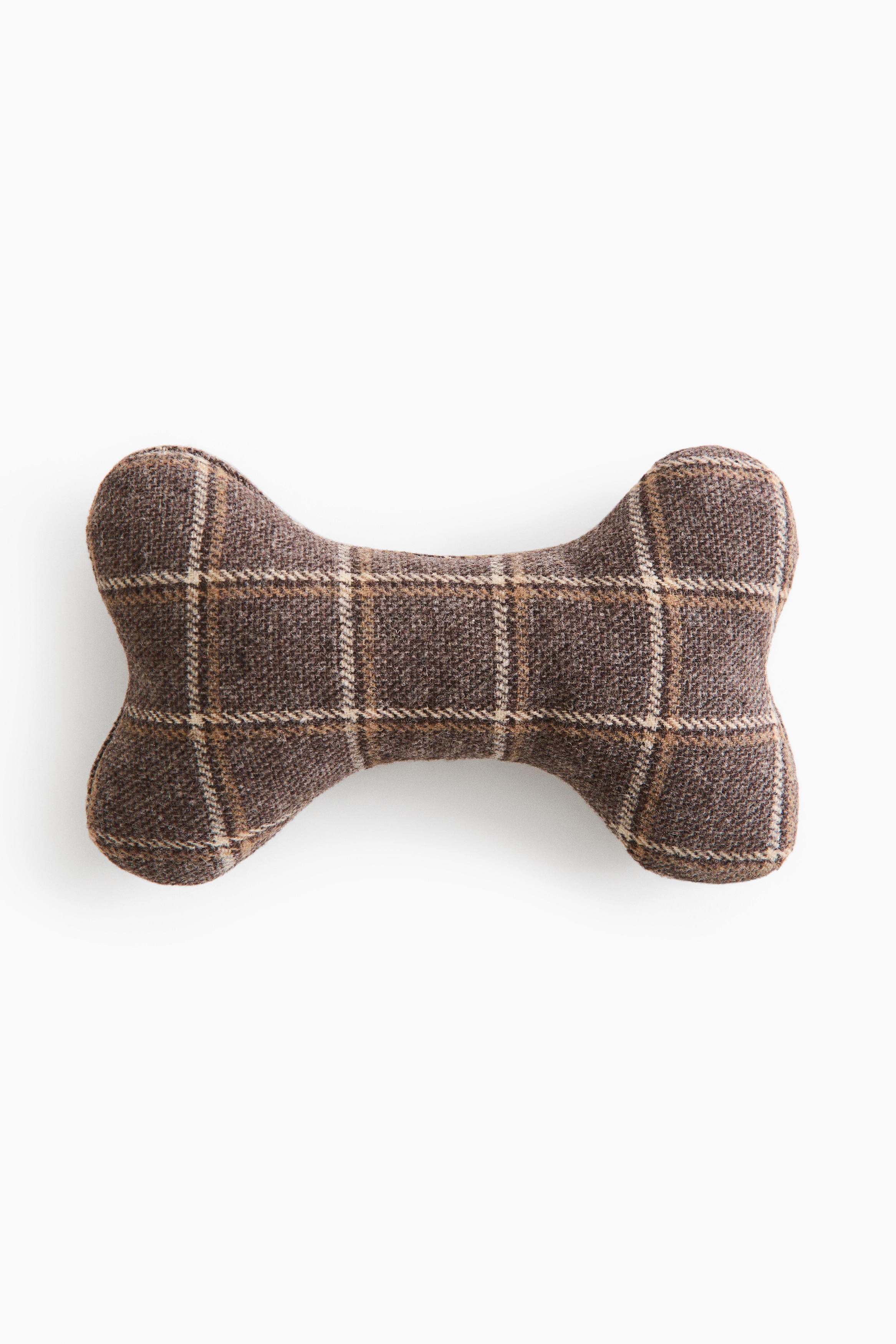 A padded, bone-shaped pet toy made from a dark, patterned woven fabric is resting on the floor in front of a dog’s paws as it lies down. The toy features a rounded, stuffed structure with visible stitched seams.