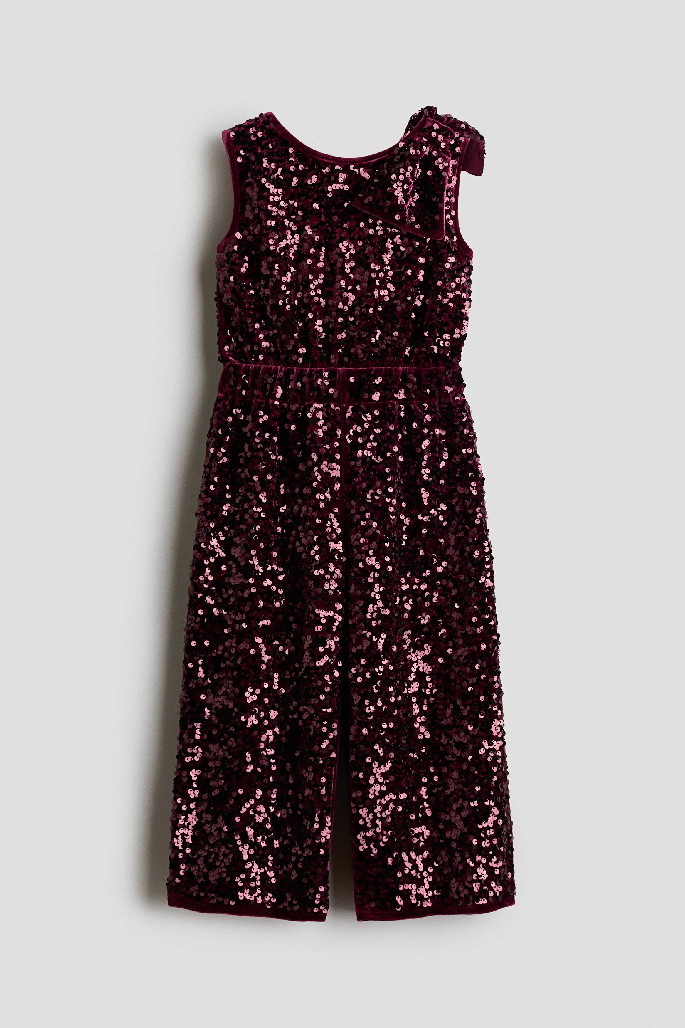 A sleeveless burgundy playsuit in sequin-covered velour is shown from a side profile facing right, showcasing its shimmering texture and velour-trimmed armhole. A matching sequined drawstring bag is hanging over the shoulder, coordinating with the playsuit.