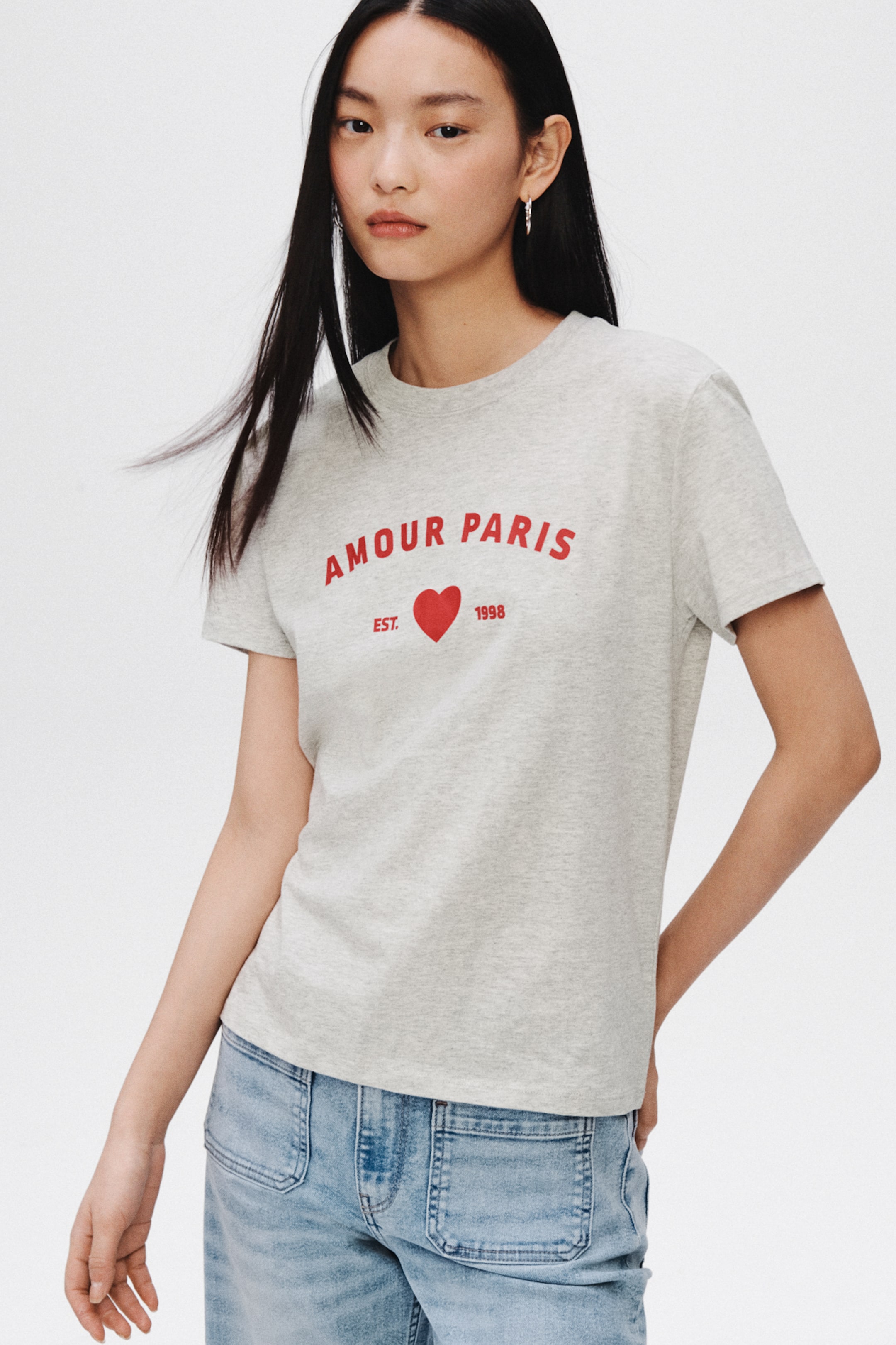View larger image: A heather gray crew neck T-shirt displays a red chest graphic featuring "AMOUR PARIS" arched above a red heart and "EST. 1998." Short sleeves and a regular fit are visible from a slight front-left angle.