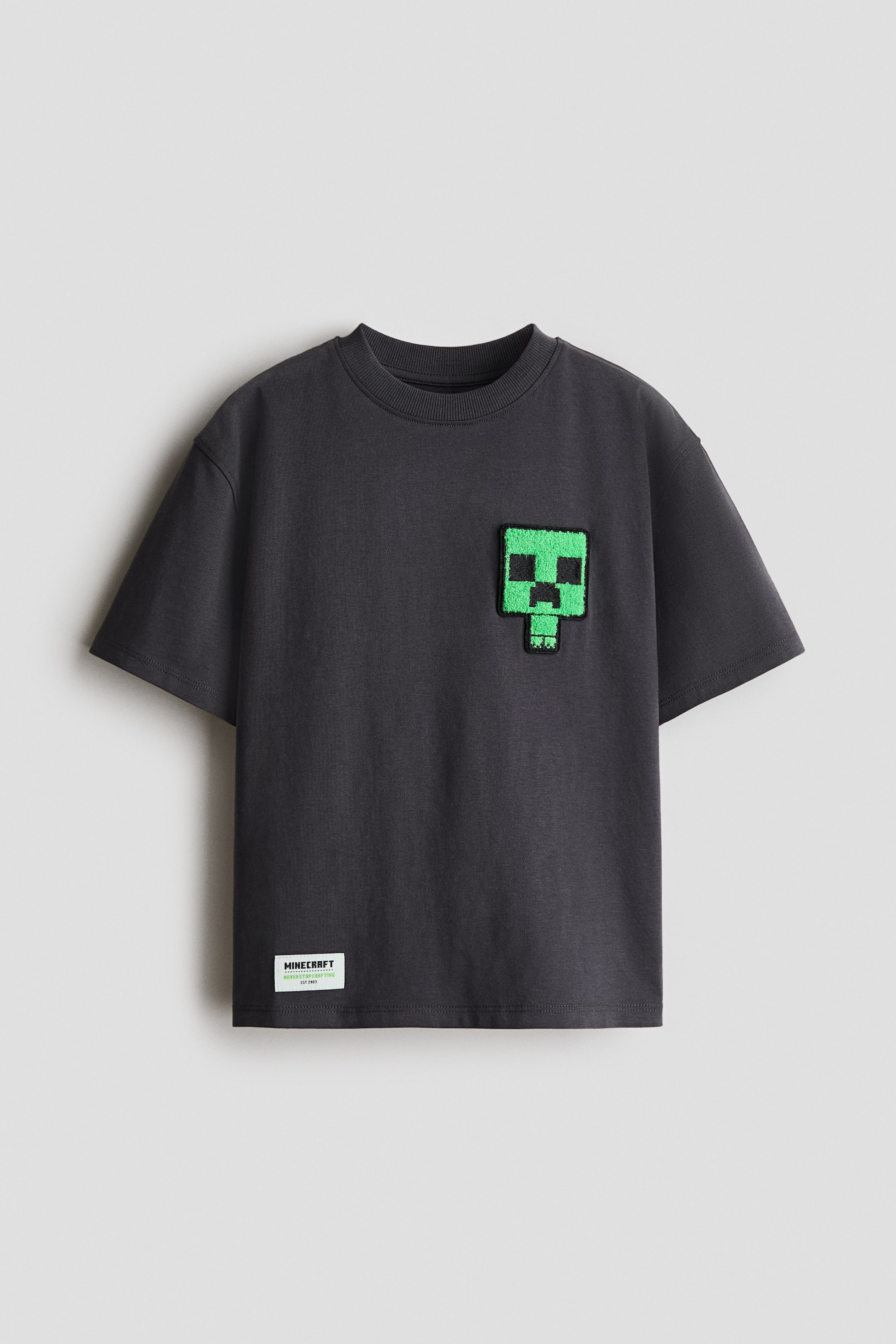 Visa större bild: En överdimensionerad t-shirt i mörkgrå mjuk bomullsjersey presenteras framifrån, med ett texturerat grönt, pixelerat ansiktsmotiv på framsidan, en rund halsringning med ribbkant och nedhasade axlar. En vit 'Minecraft'-etikett är synlig nära den nedre vänstra fållen.