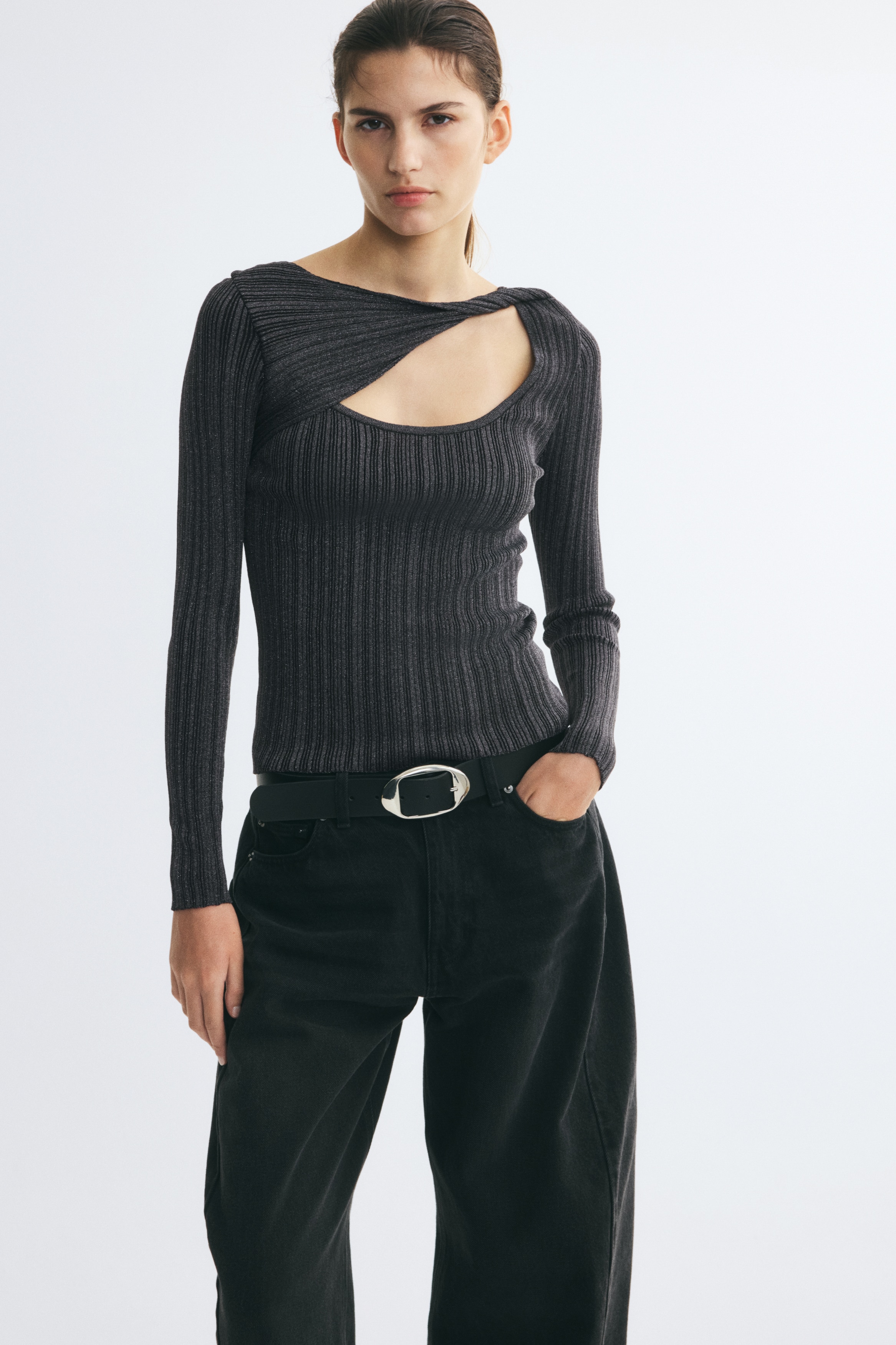 Un top a maniche lunghe in maglia a coste, grigio scuro e metallizzato, Ã¨ mostrato frontalmente, con una silhouette aderente, coste verticali e maniche lunghe. Il capo presenta uno scollo asimmetrico, con un inserto diagonale che crea unâapertura a goccia sopra uno scollo tondo.