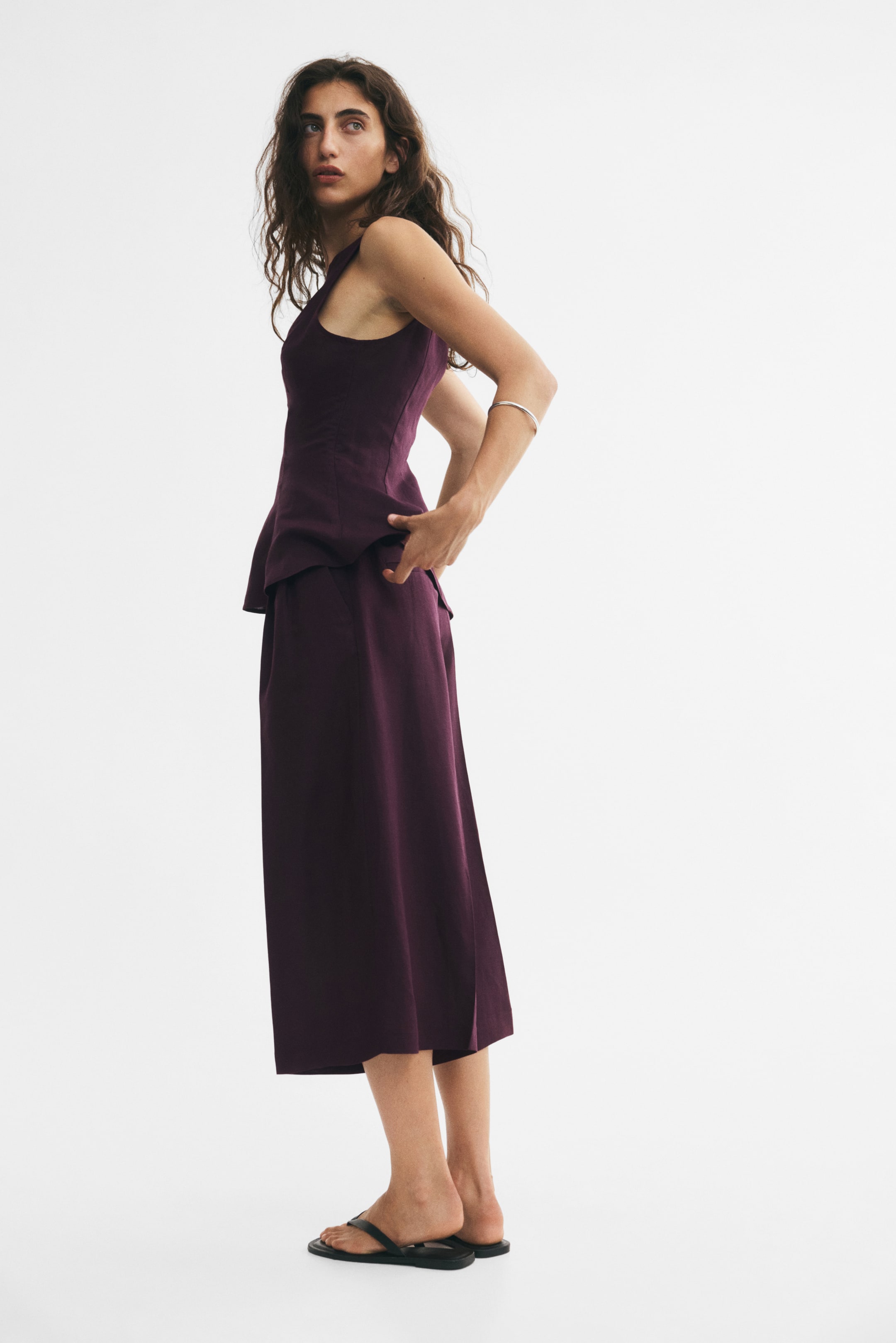 Ampliar la imagen: Unos culottes de corte holgado en color morado profundo, confeccionados en una mezcla de lino, son llevados por la modelo; presentan piernas amplias con pinzas en la parte superior y pliegues marcados. La modelo coloca las manos en la cintura de los culottes, cerca de los bolsillos laterales diagonales, y se combinan con un top sin mangas a juego.