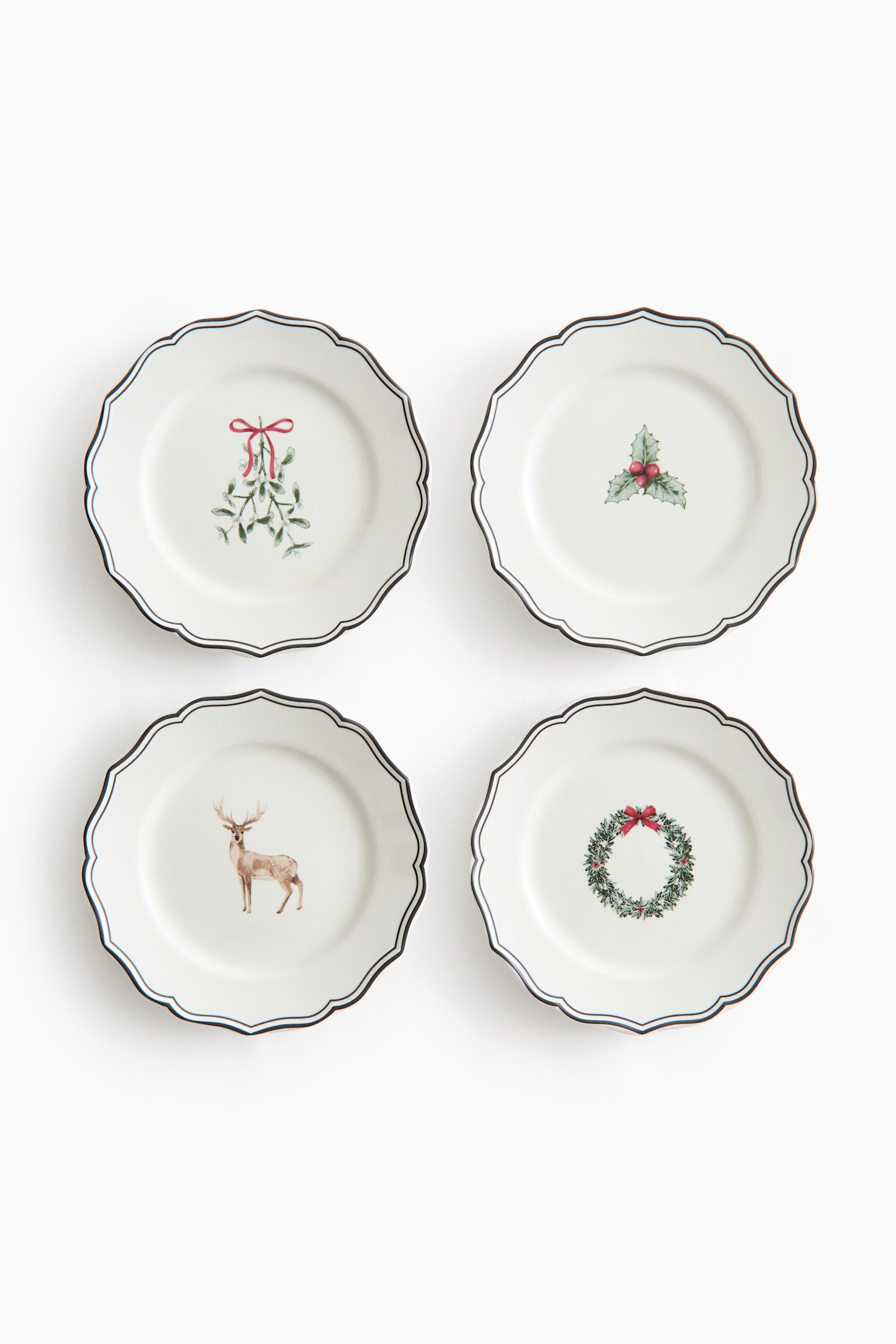 Four white ceramic plates are shown from the front, each featuring a scalloped edge with a thin, dark double border. Each plate displays a different central illustration, including mistletoe tied with a red ribbon, a holly sprig with red berries, a standing deer, and a green wreath with a red bow. - 1