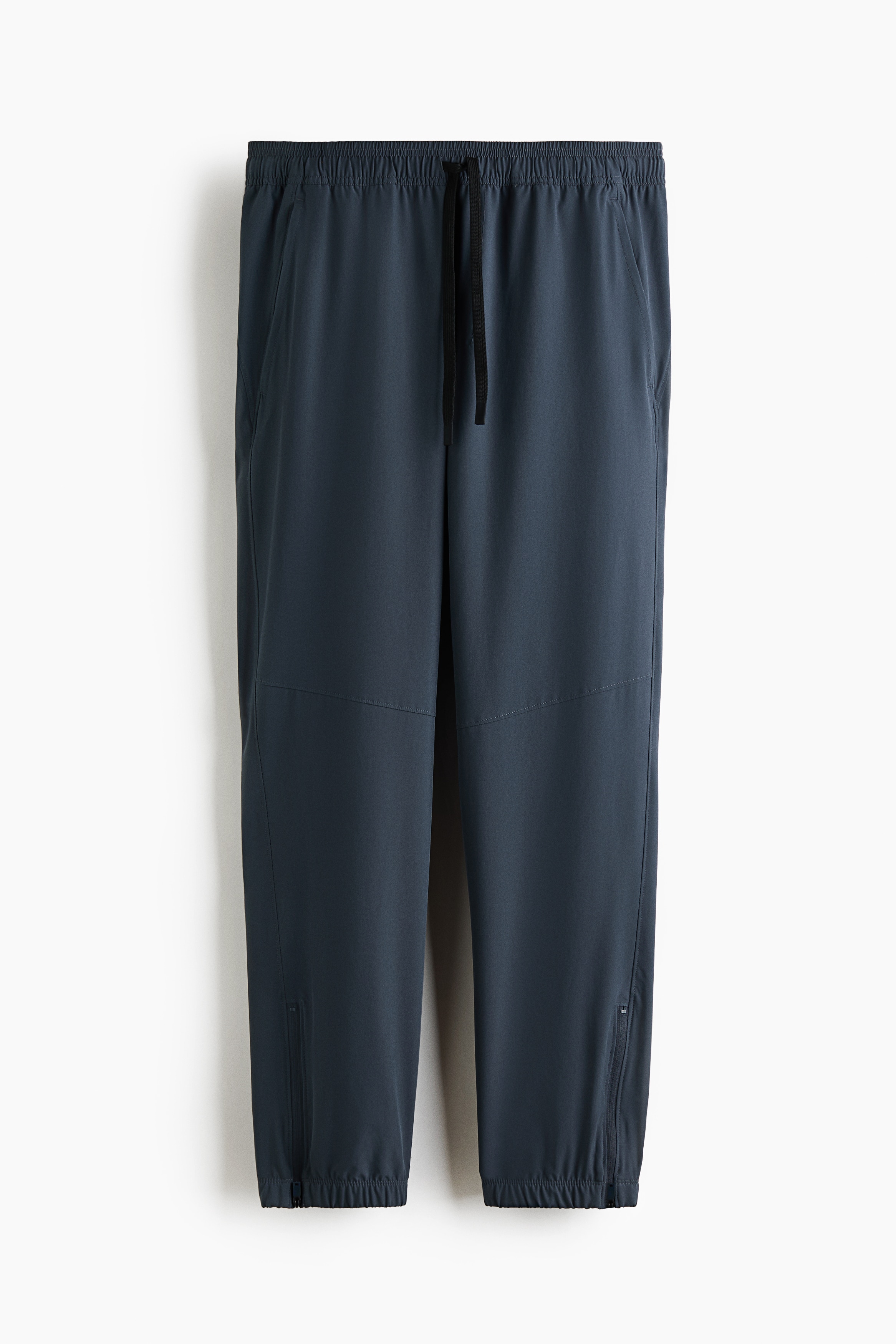 Model nosi tamnosmeÄu majicu dugih rukava sa lažnim rol-kragnom i sportske pantalone u Å¡kriljasto plavoj boji. Pantalone imaju elastiÄne manžetne sa detaljima na rajsferÅ¡lus, a autfit je stilizovan svetlosivim patikama i svetlosivom kapom noÅ¡enom unazad.