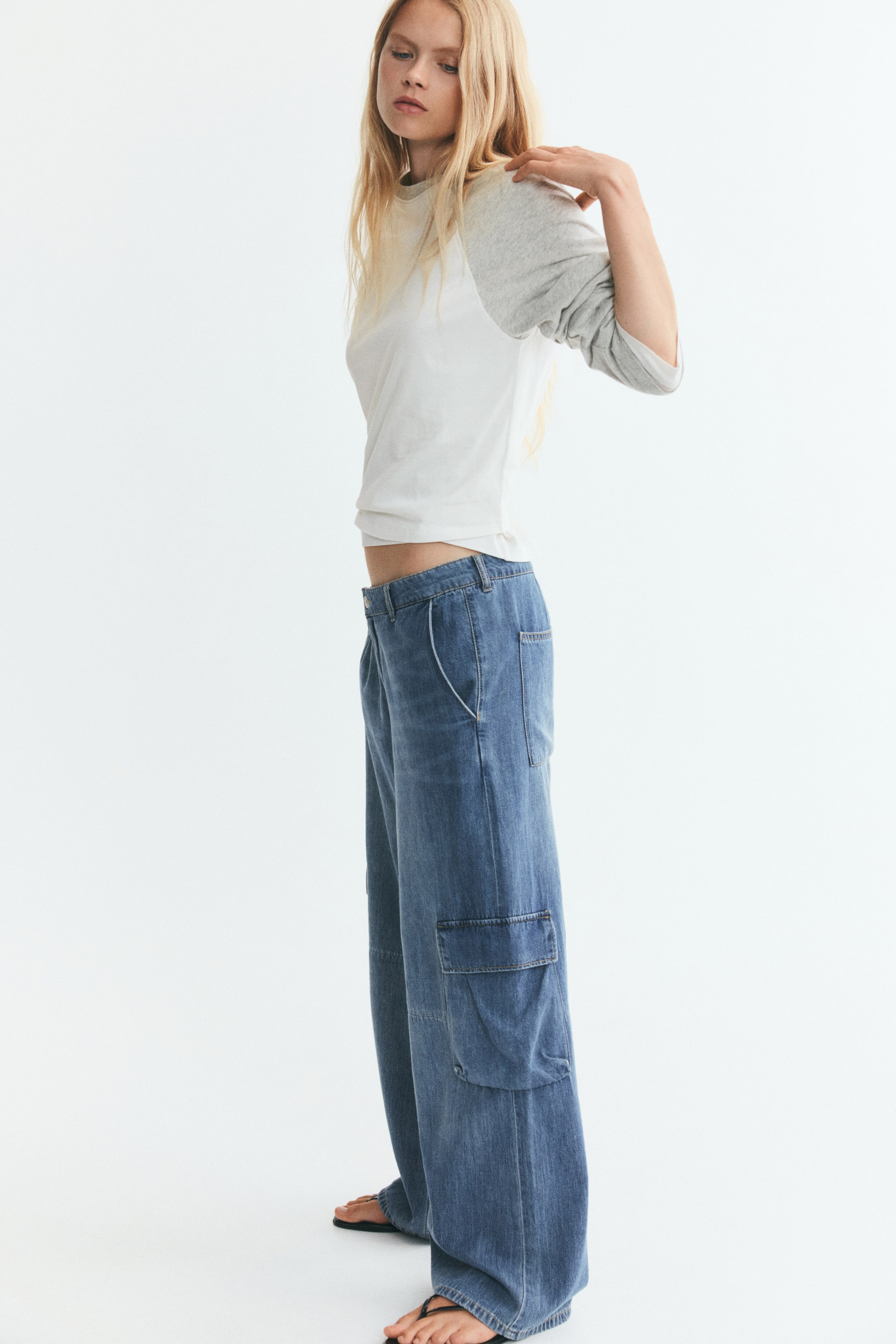 Visualizza immagine più grande: Un top in jersey di cotone bicolore, con corpo bianco, scollo tondo e maniche lunghe raglan grigio chiaro, indossato dalla modella. Il top è abbinato a jeans cargo blu a gamba larga, mentre la modella alza una mano alla spalla, arricciando leggermente la manica.