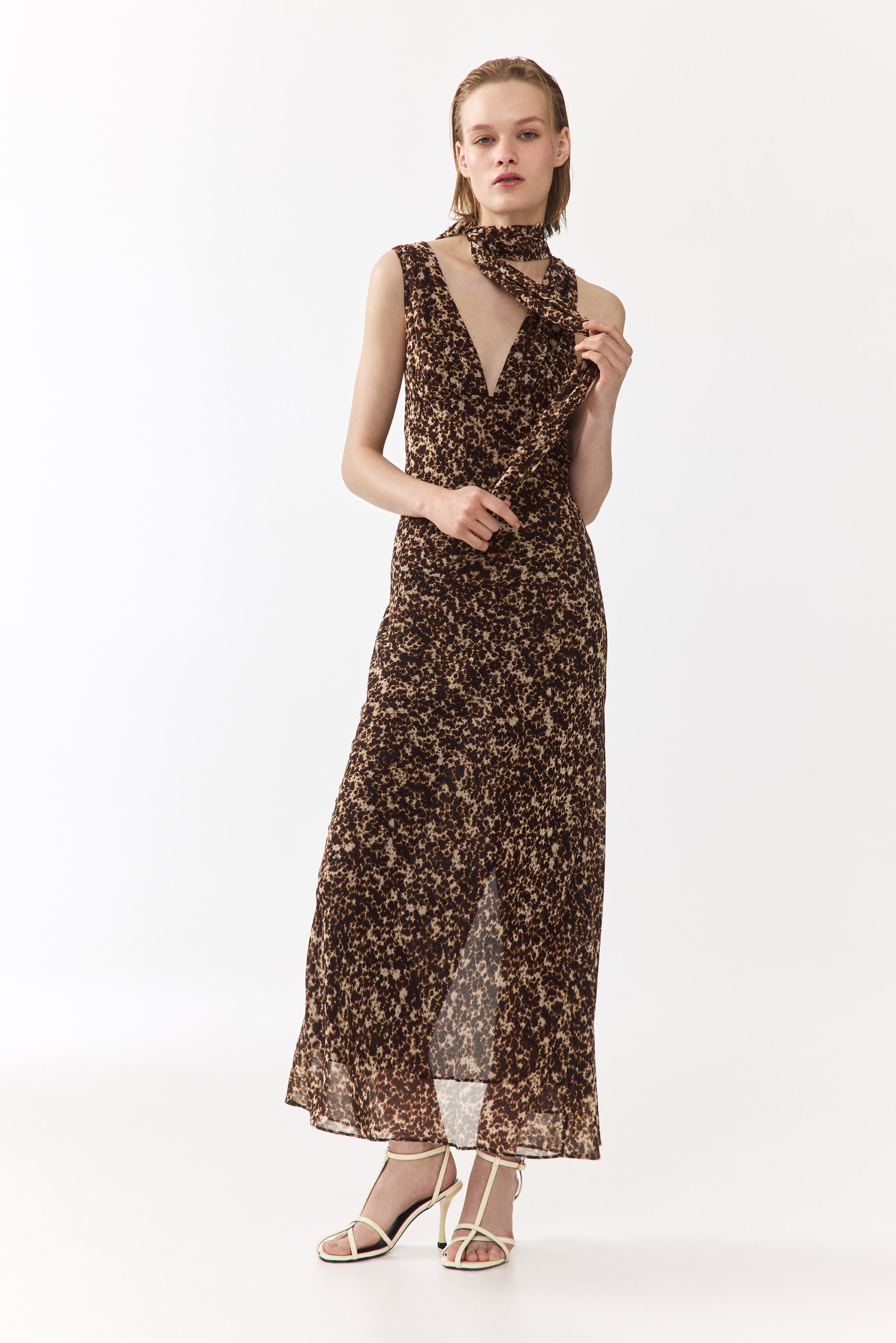 Ver imagen más grande: Un vestido con un estampado abstracto en tonos marrón y beige, un escote en V profundo y un cuerpo sin mangas. Está diseñado con una falda de longitud maxi, una abertura alta en la parte delantera y una bufanda a juego atada alrededor del cuello, orientada hacia adelante.