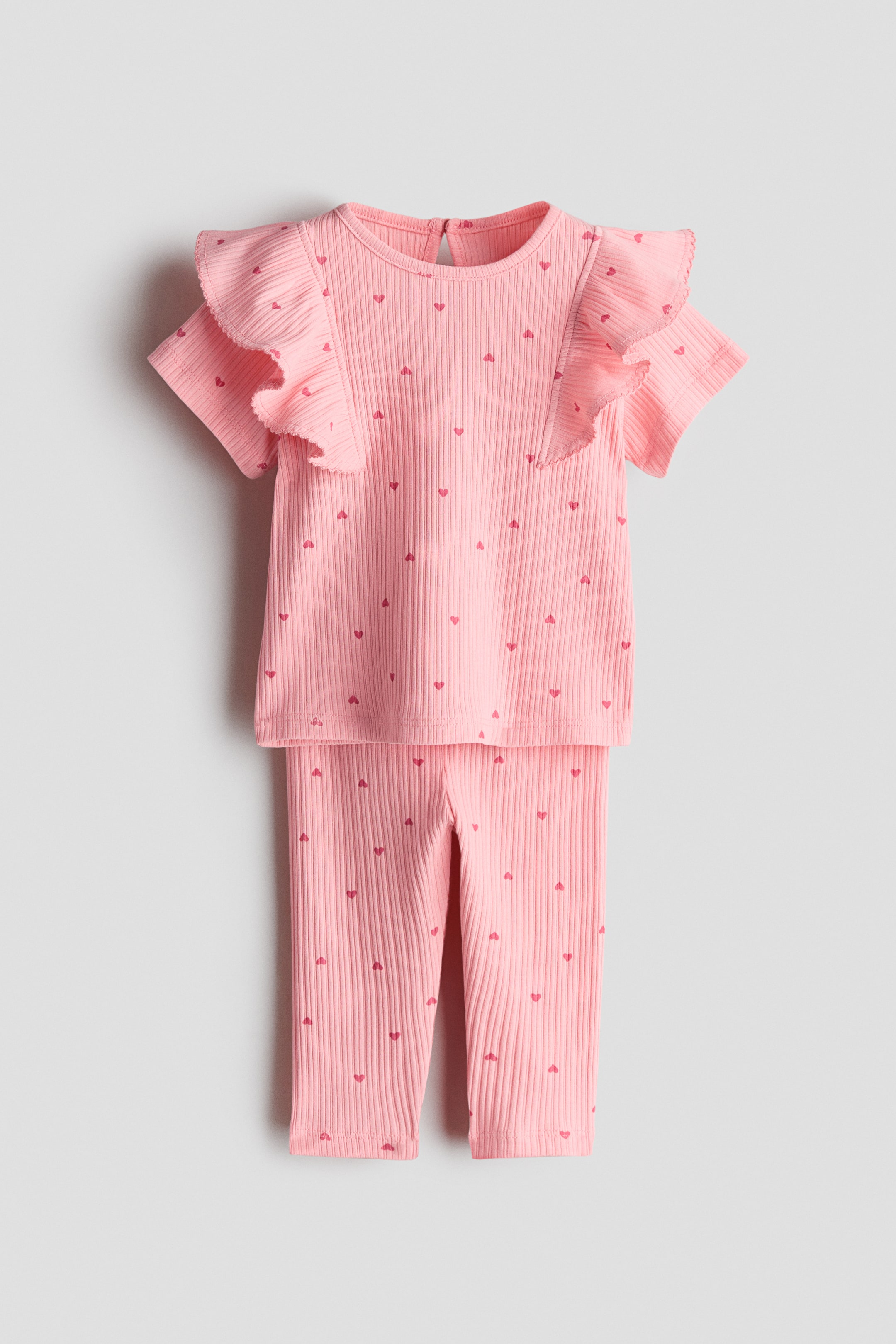 View larger image: A pink ribbed jersey top and leggings set is shown from the front. The short-sleeved top features wide, picot-trimmed frills at the shoulders and a round neckline, while both pieces display a repeating pattern of small red hearts.