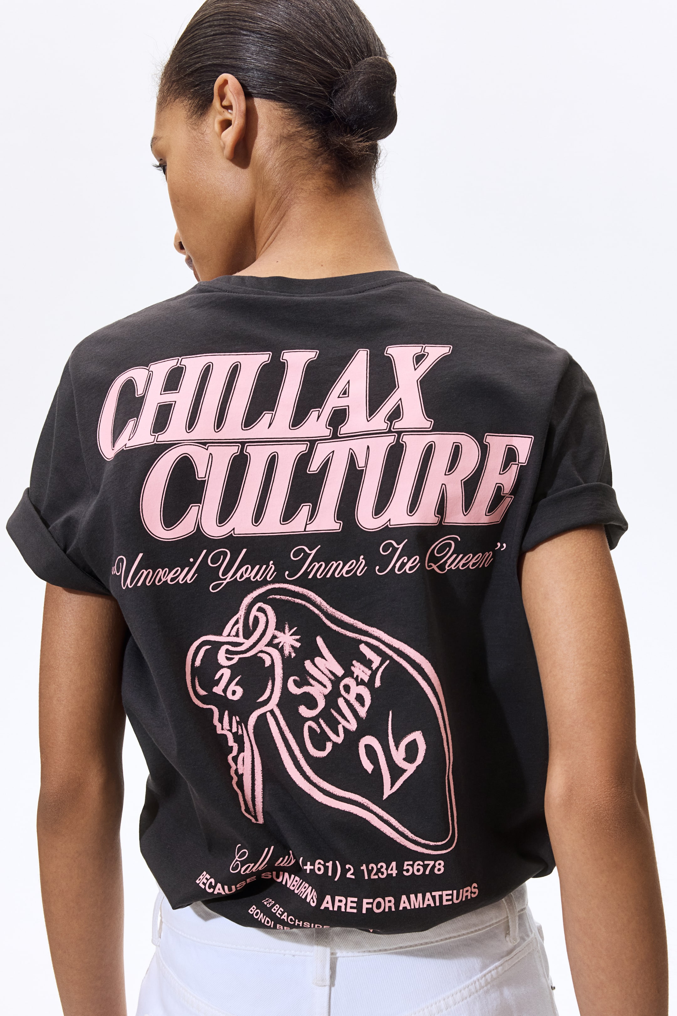 View larger image: A black t-shirt is presenting its back, featuring a large pink graphic that is displaying the prominent text 'CHILLAX CULTURE' and 'Unveil Your Inner Ice Queen'. The graphic is also showing a key design labeled 'SUN CLUB #1 26', a phone number, the phrase 'BECAUSE SUNBURNS ARE FOR AMATEURS', and location details. Its short sleeves are cuffed, and the shirt is resting above white pants.