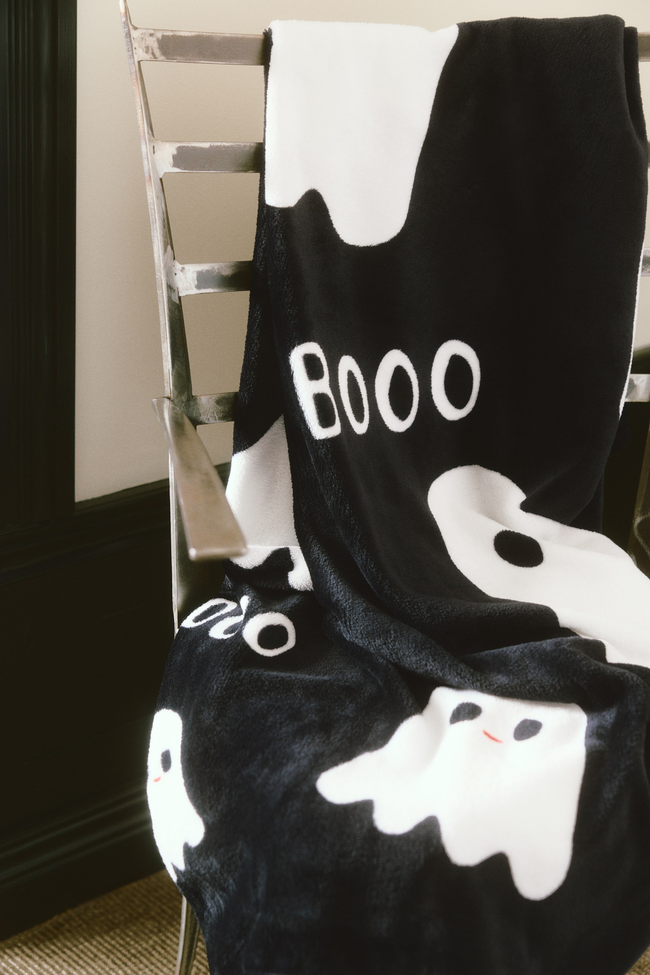 View larger image: A soft fleece blanket is draped over a metal chair, showcasing a Halloween-themed pattern of friendly white ghosts and the word "Booo" in white block letters against a black background. The plush blanket features several ghosts with tiny black eyes and red smiling mouths scattered across its textured surface.