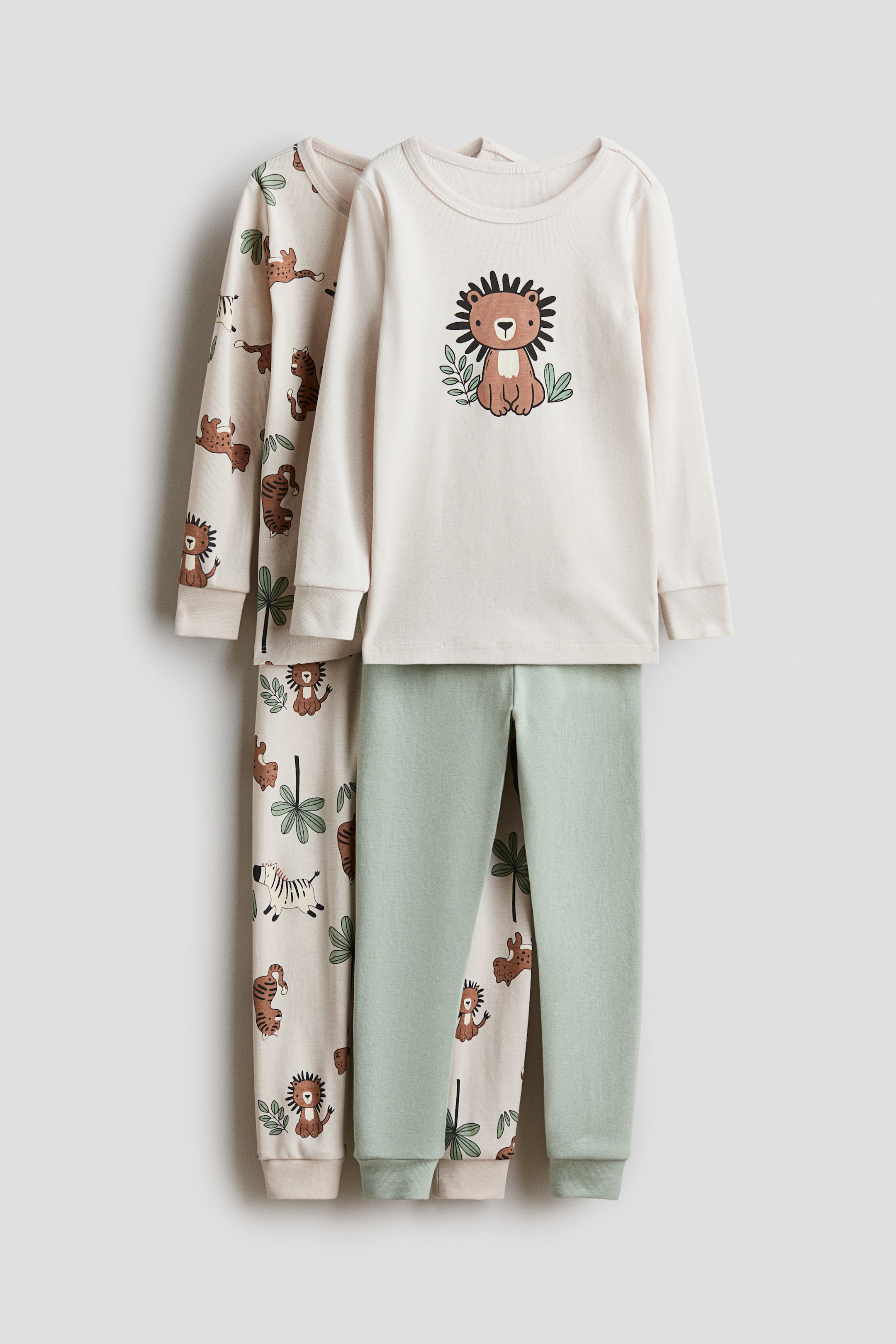 View larger image: Two sets of soft cotton jersey pajamas are being presented. The front set, facing front, includes a long-sleeved cream top with ribbing at the neckline and cuffs, featuring a lion graphic, paired with light green bottoms with an elastic waistband and ribbed hems. The second set, partially visible behind, features a cream long-sleeved top and matching bottoms with an all-over safari animal print.