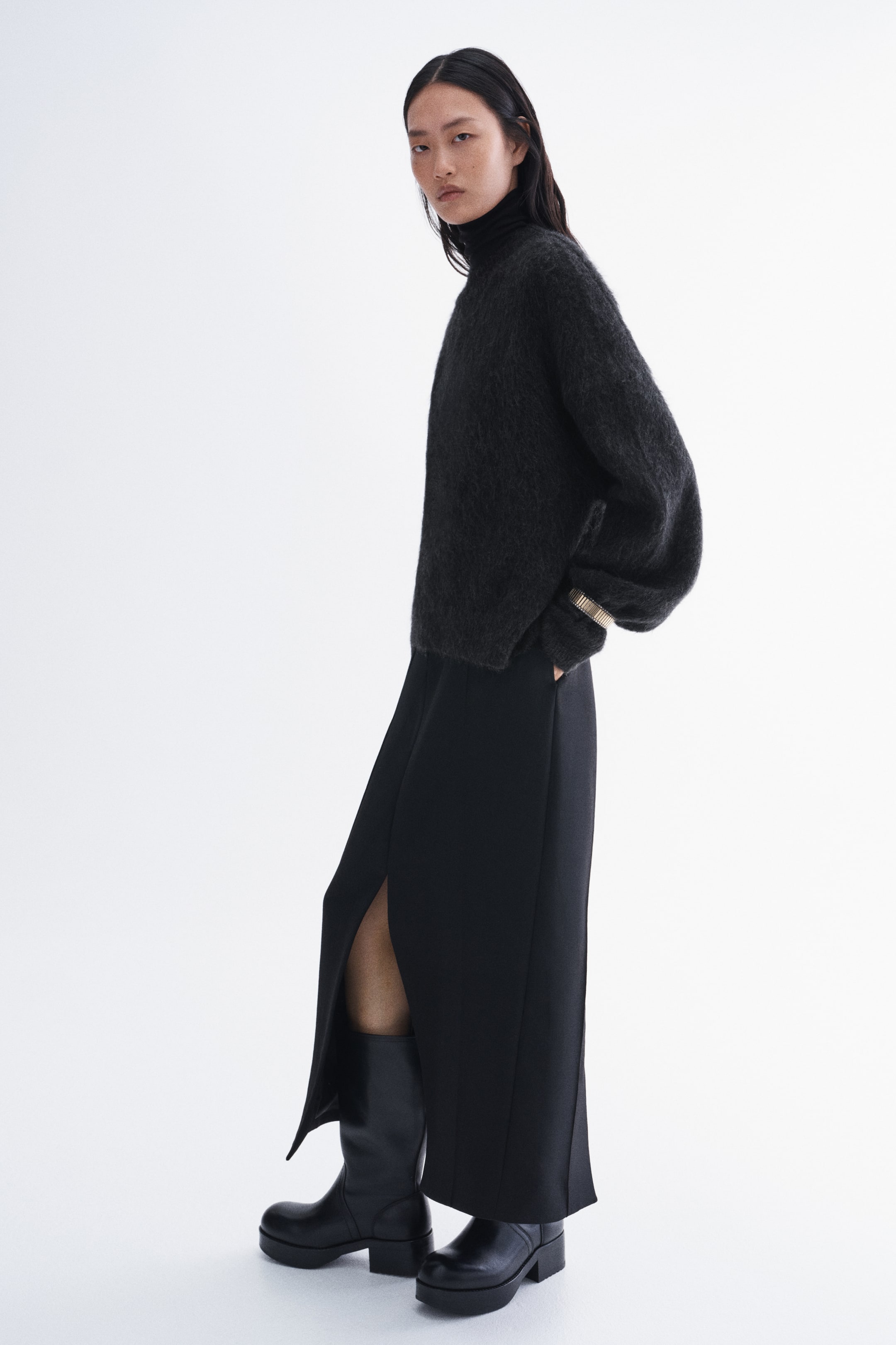 Grösseres Bild ansehen: Der dunkelgraue, flauschige Strick-Rollkragenpullover wird vom Model getragen und zeichnet sich durch eine Oversize-Passform und lange Ärmel aus. Der Pullover ist über einem schwarzen Midirock mit hohem Schlitz und schwarzen Plateau-Stiefeln gestylt, wobei am Handgelenk des Models ein goldfarbener, gerippter Armreif zu sehen ist.