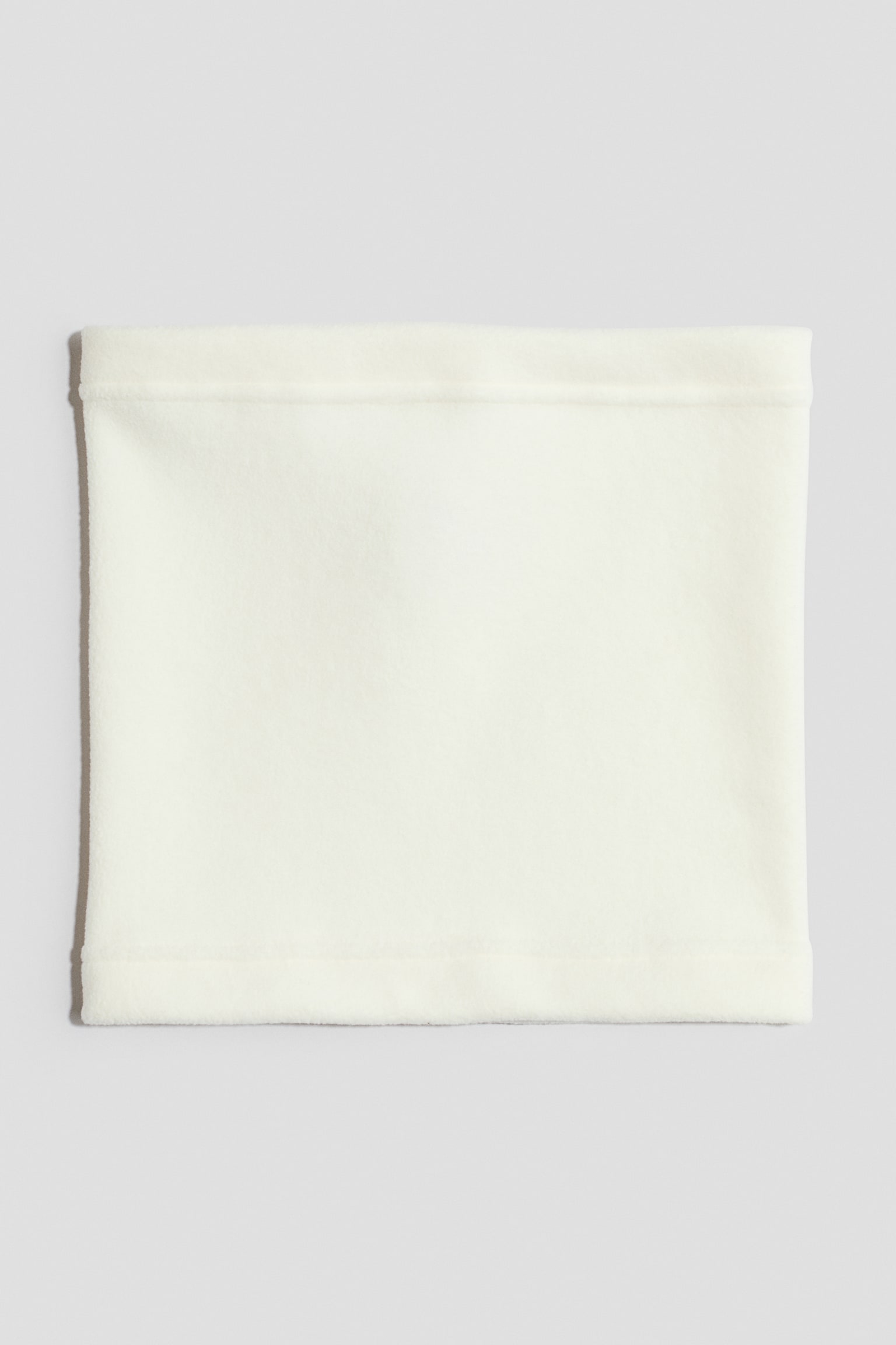 Una bufanda tipo tubo de tela polar color blanco roto estÃ¡ extendida sobre una superficie plana, mostrando una vista frontal de su forma rectangular y su textura suave. La bufanda presenta dobladillos terminados y cosidos en las aberturas superior e inferior.
