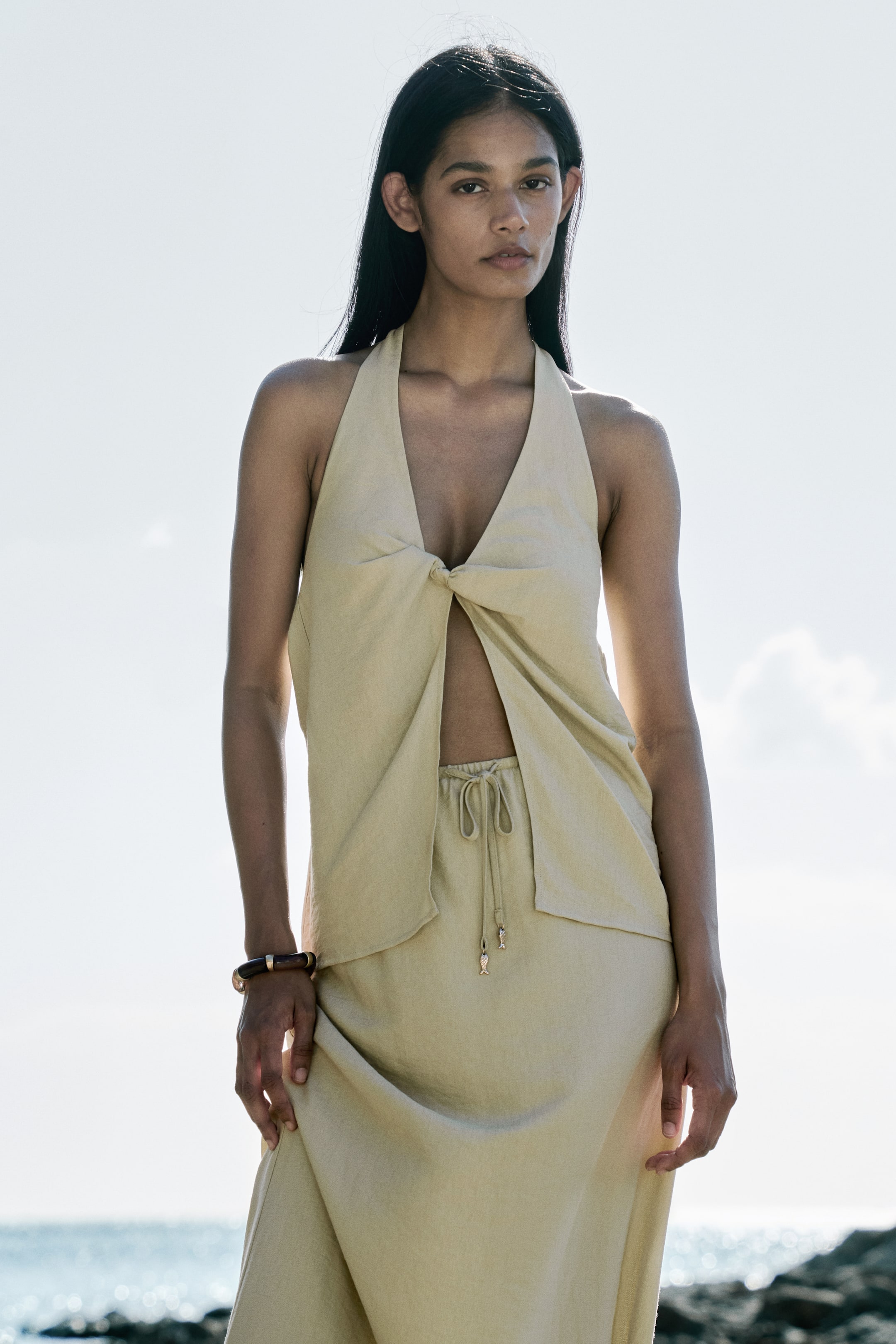 View larger image: Light beige two-piece ensemble featuring a halter top with a twisted knot at the centre front, creating a deep V-neckline with an opening below the bust. The matching long skirt showcases a drawstring waist detailed with two small fish-shaped charms.