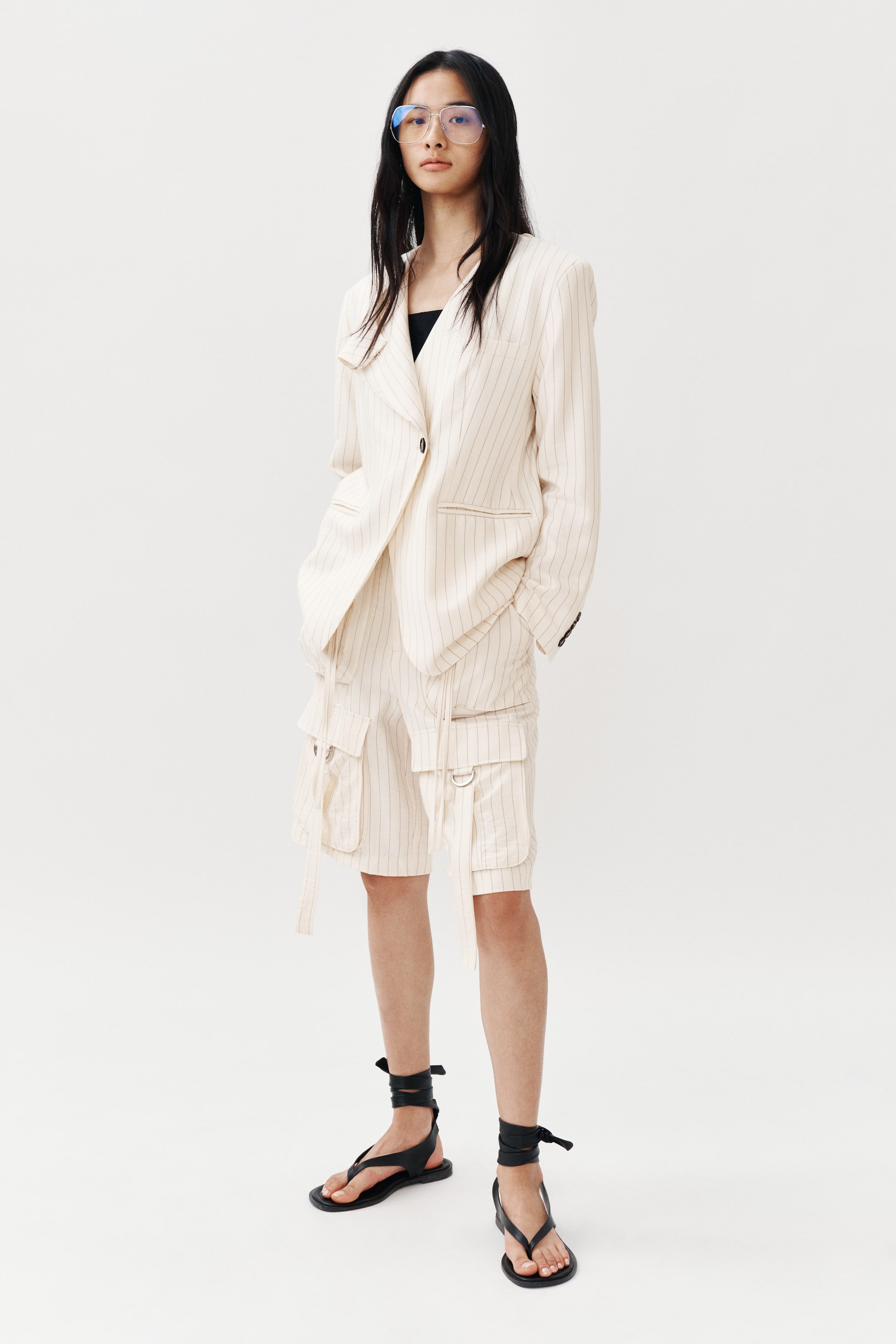 View larger image: "Sebuah blazer pinstripe warna putih pudar dan seluar pendek kargo yang sepadan sedang dipamerkan, menghadap ke depan. Blazer bersaiz besar ini menampilkan lapel berlekuk, penutupan satu butang gelap, poket dada, dan dua poket flap, dan dipakai di atas baju hitam. Seluar pendek kargo ini mempunyai pelbagai poket sisi dengan tali gantung dan cincin D."