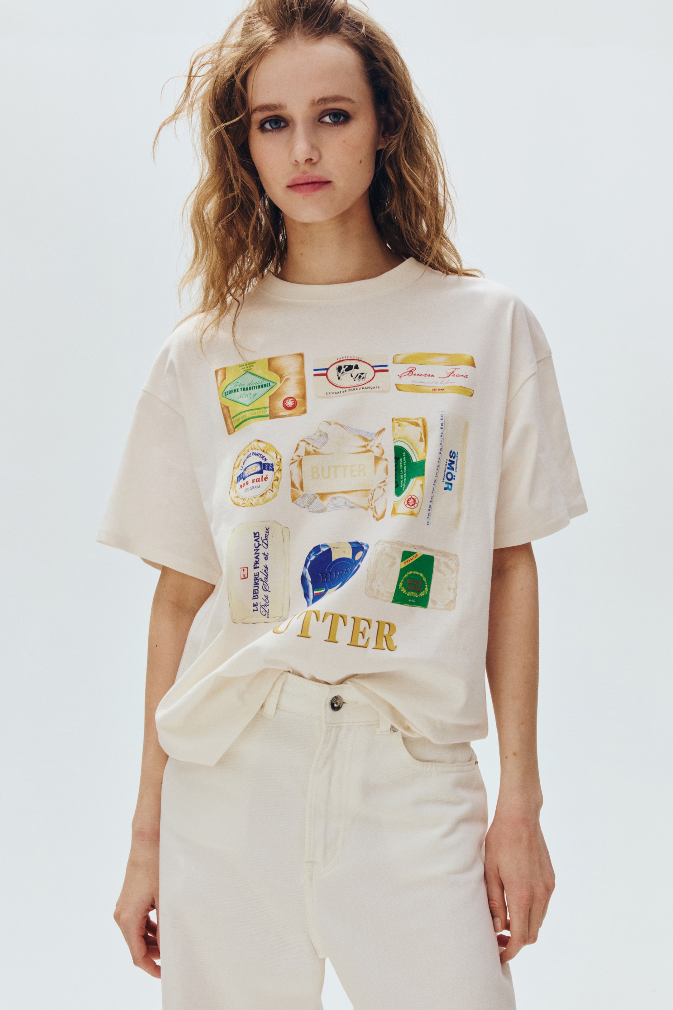 Agrandir l'image: Un t-shirt oversize en jersey de coton doux, arborant un motif imprimé de divers designs d'emballages de beurre sur le devant, est porté par le modèle. Il présente un col rond côtelé, des épaules tombantes et un ourlet droit qui est rentré dans un pantalon de couleur claire.