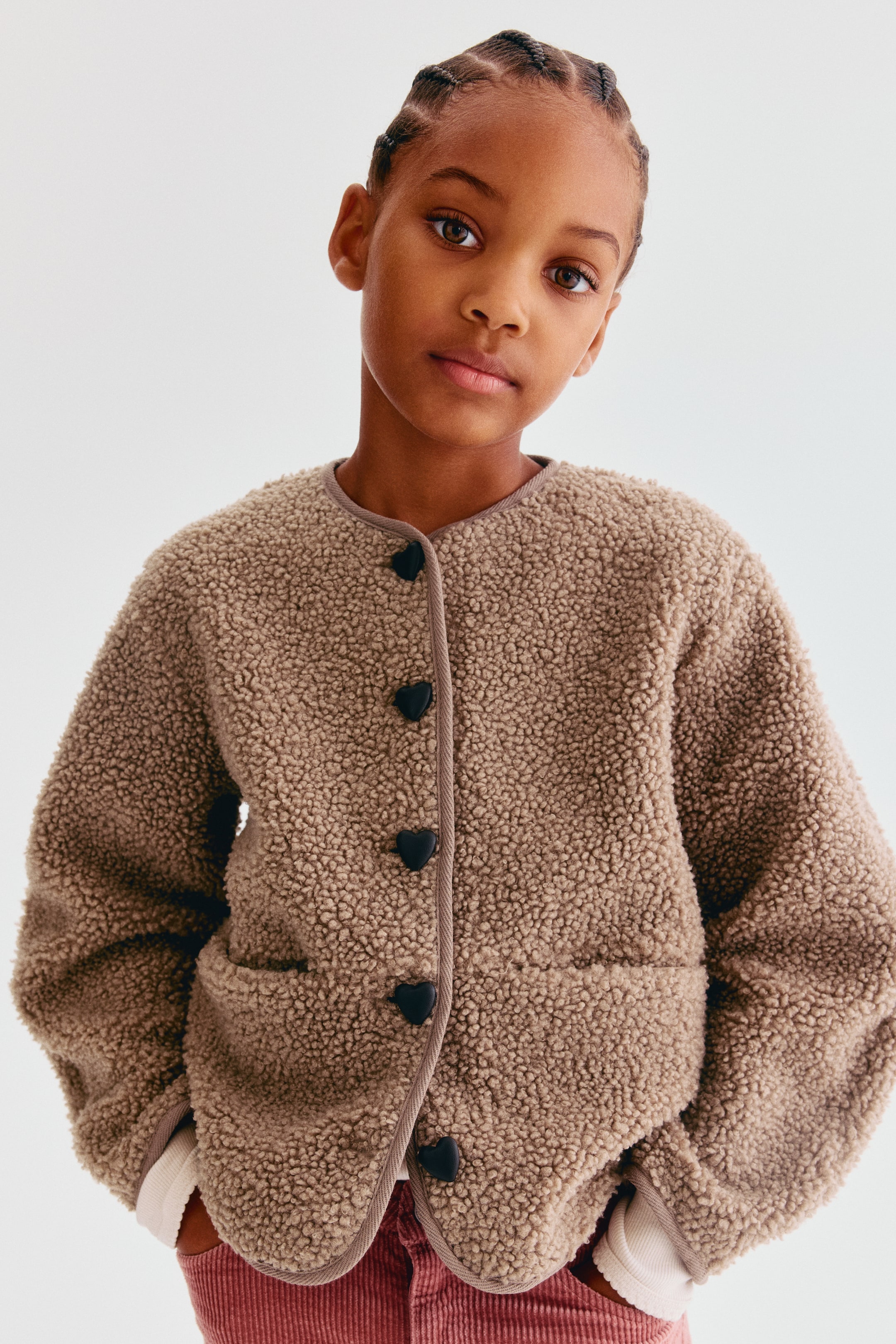 Voir une image plus grande: Une veste teddy beige est montrée de face, avec des épaules tombantes, un col sans col et une ganse en sergé étroite le long des bords. La veste se ferme sur le devant avec des pressions noires en forme de cœur et présente des poches passepoilées discrètes à l’avant.