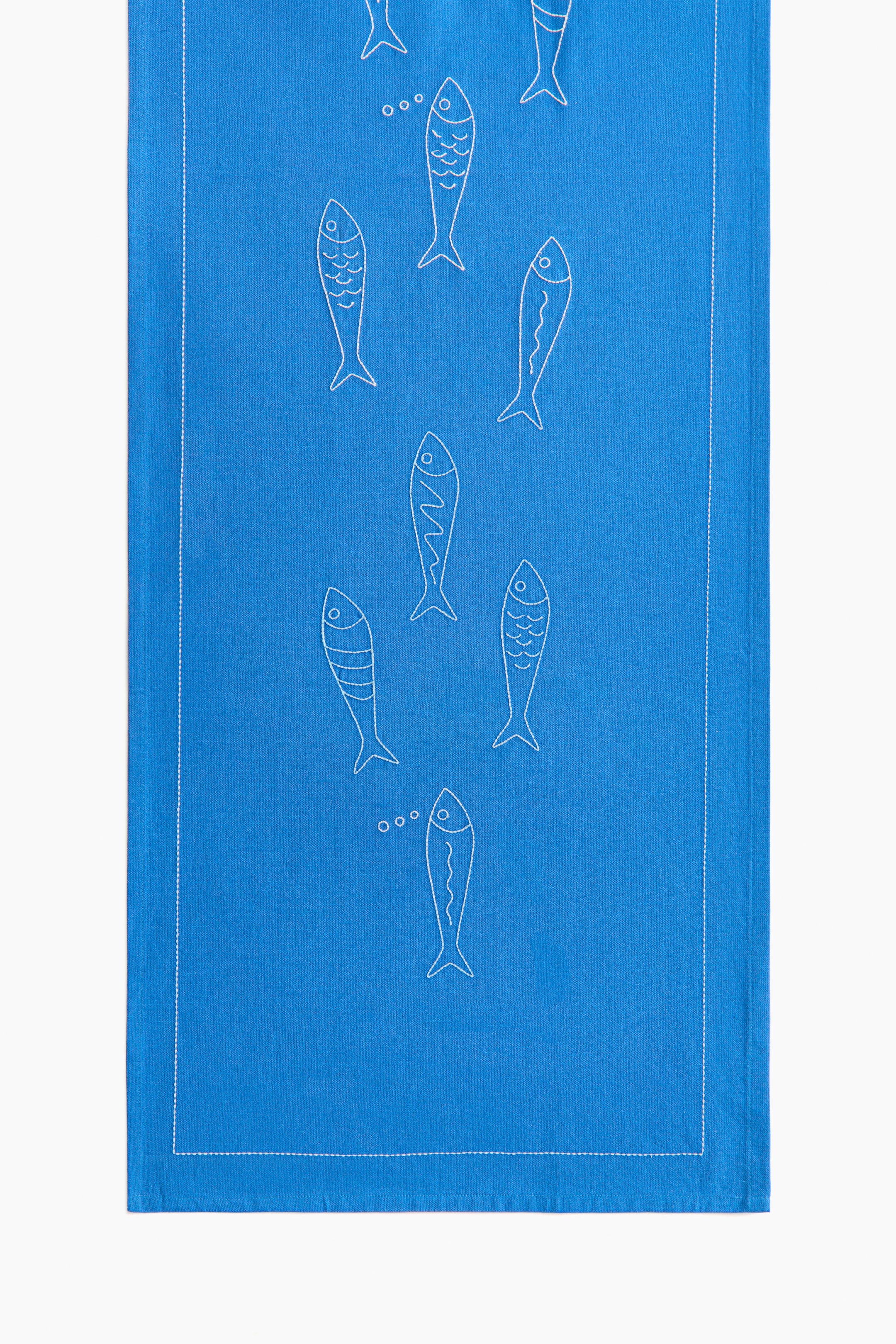 Ver imagen más grande: Un camino de mesa azul, confeccionado con una mezcla de algodón y lino, presenta diseños de peces bordados en blanco a lo largo de su parte frontal. El borde está delineado con costuras blancas, y se remata con un dobladillo plegado de 1 cm.
