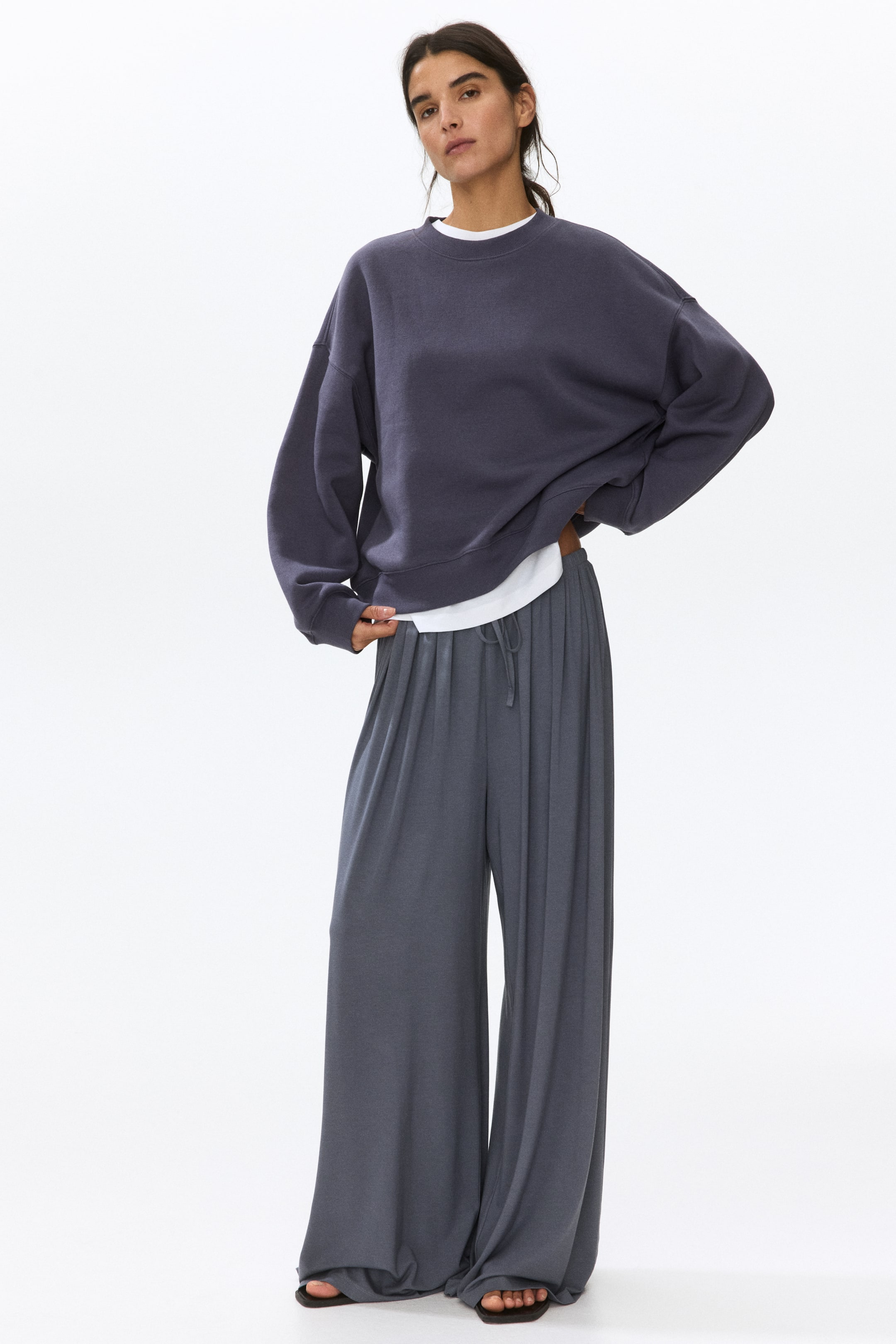 Voir une image plus grande: Un chandail à col rond surdimensionné gris foncé, avec des poignets et un ourlet côtelés, est superposé à un t-shirt blanc. Cela s'accompagne d'un pantalon ample à cordon de serrage gris foncé, qui tombe sur des sandales plates foncées.