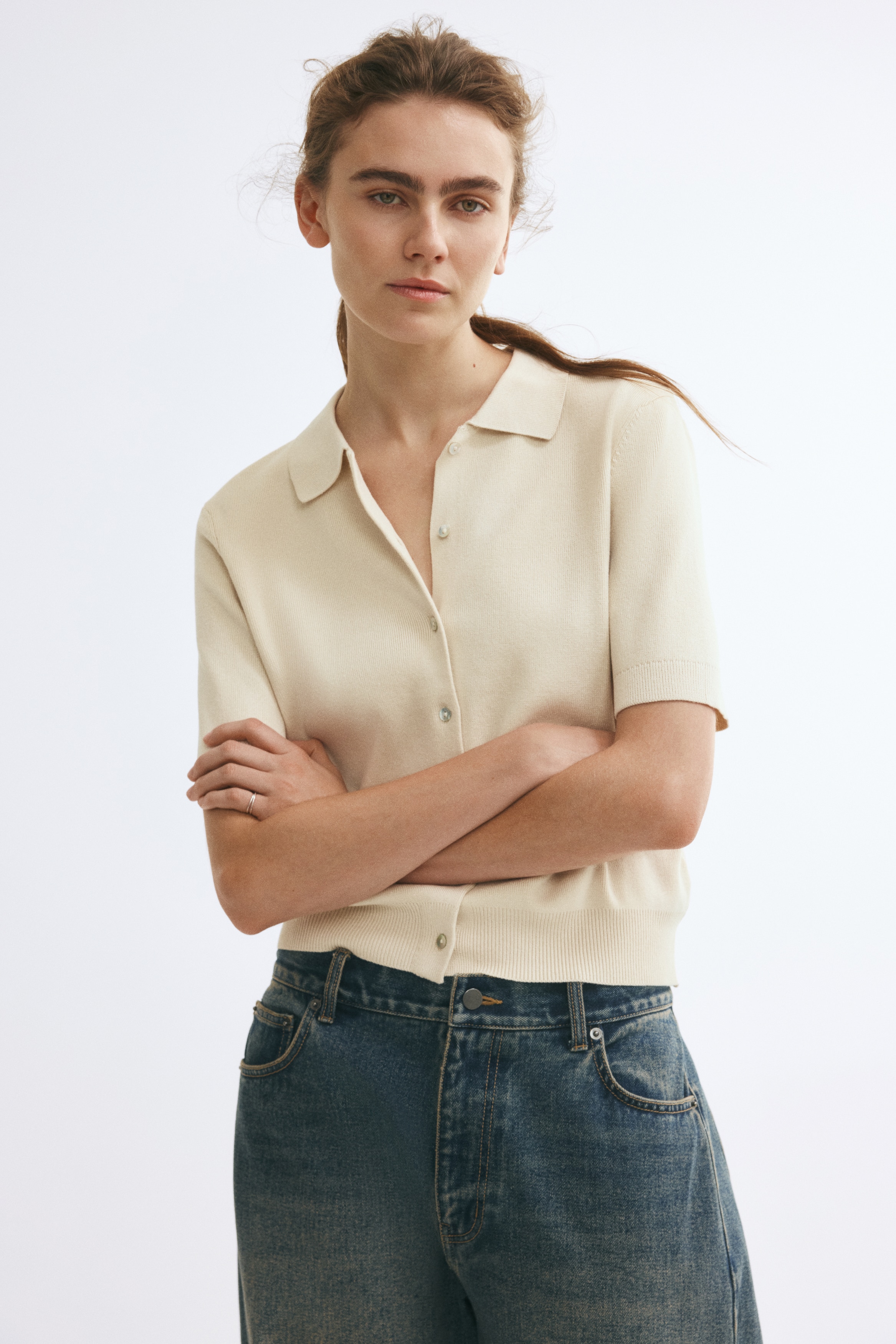 A cream-coloured, short-sleeve knit polo cardigan is facing front, featuring a flat-knit collar, a full-length button-down placket, and ribbed detailing at the cuffs and hem.