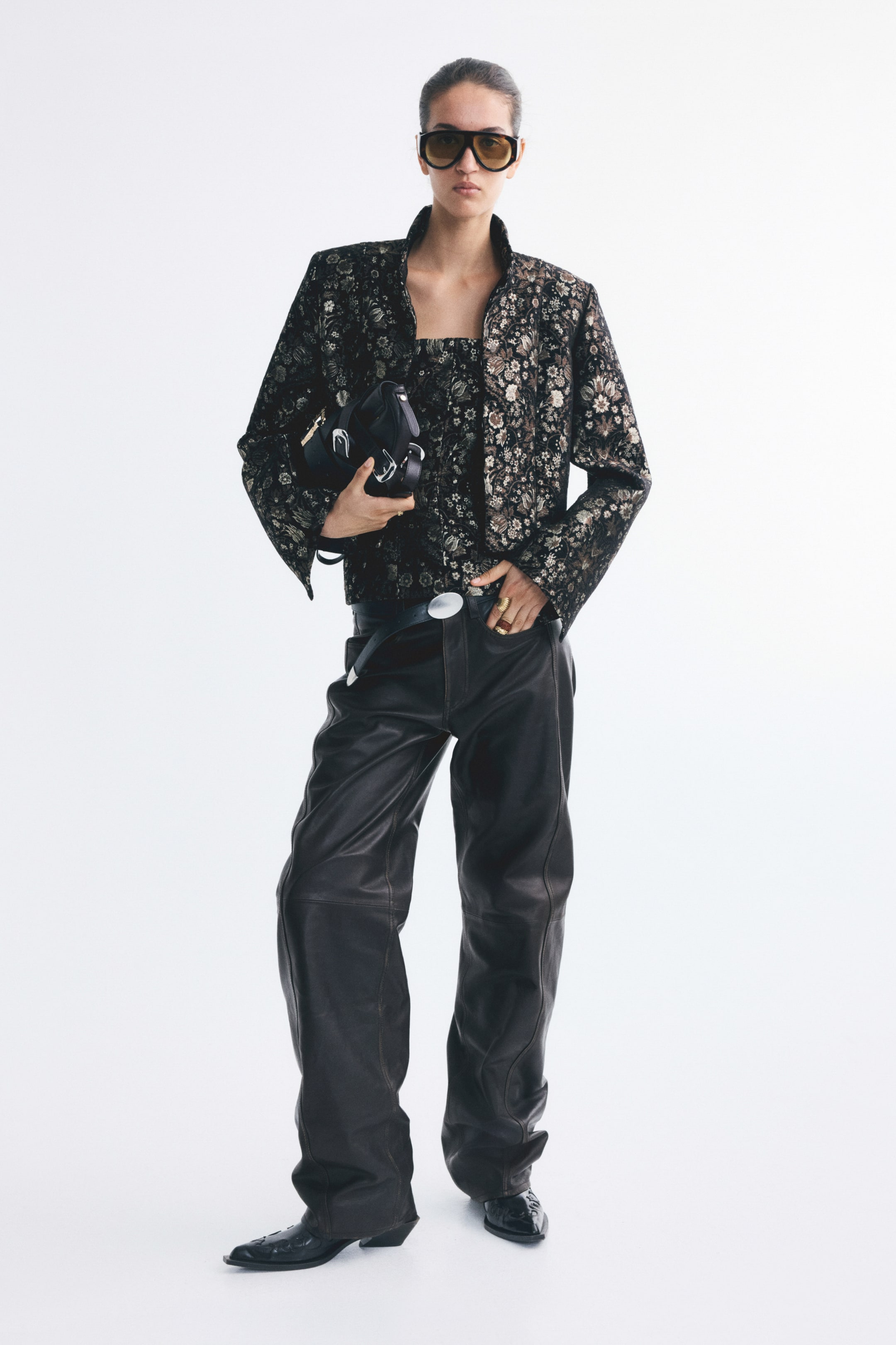 View larger image: Relaxed-fit leather trousers with a barrel-leg silhouette and prominent shaping seams are worn by the model, styled with a black leather belt. The model is standing with one hand resting in the front pocket of the trousers, which are paired with a dark floral jacket and black pointed-toe boots.