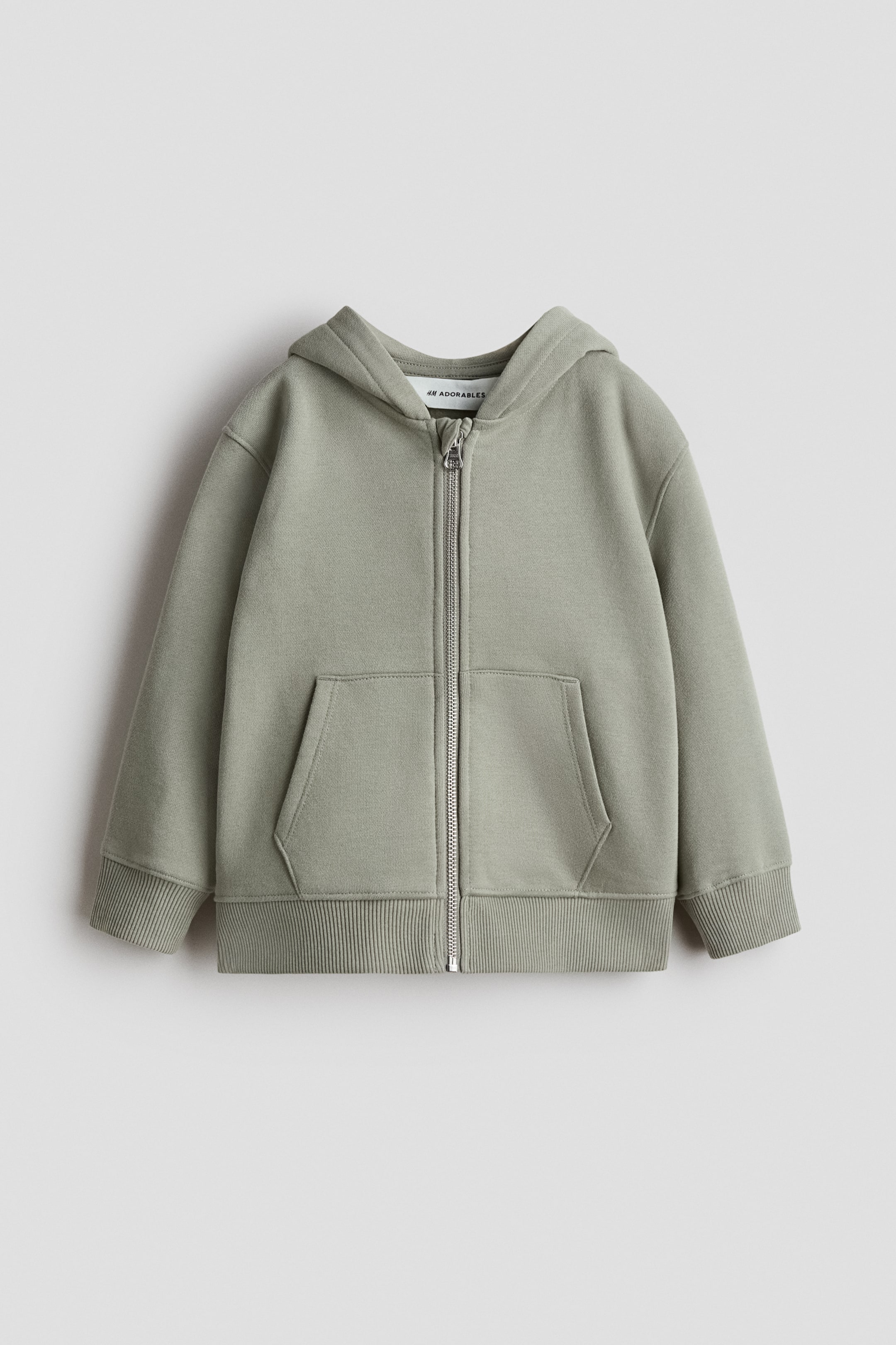 Ingrandisci l'immagine: Un H&M Adorables felpa con zip in verde salvia è presentata dal DAVANTI, caratterizzata da un cappuccio, una cerniera centrale e tasche a marsupio. È progettata con spalle scese, polsini a coste e un orlo a coste.