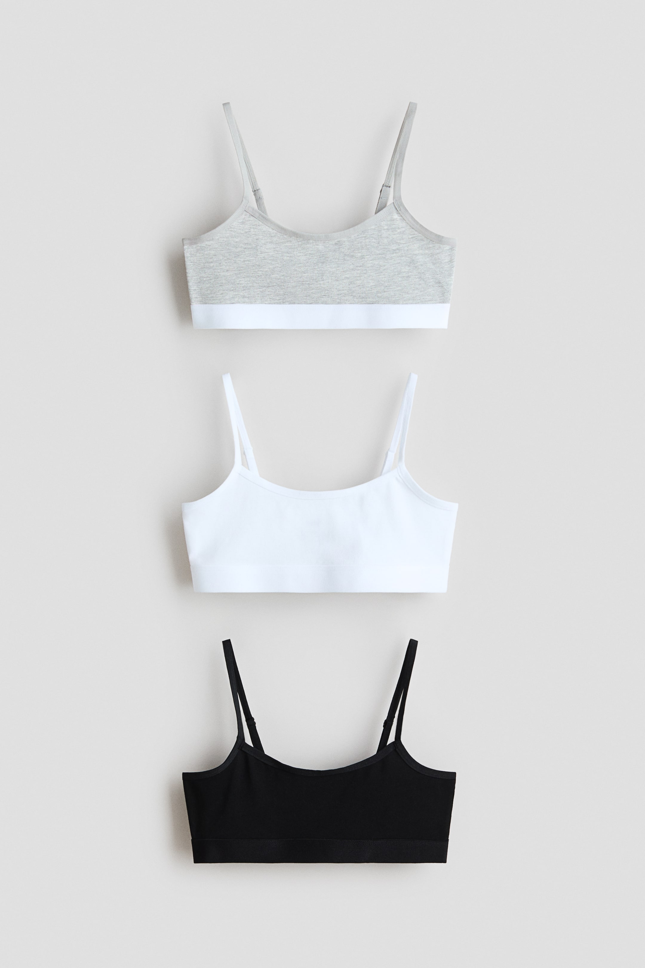 View larger image: Three front-facing bralettes are being showcased, arranged in a vertical stack. The top bralette is a light grey marl with a white elastic underband, the middle one is pure white with a matching white elastic underband, and the bottom bralette is classic black with a coordinating black elastic underband. Each piece features delicate, adjustable straps.