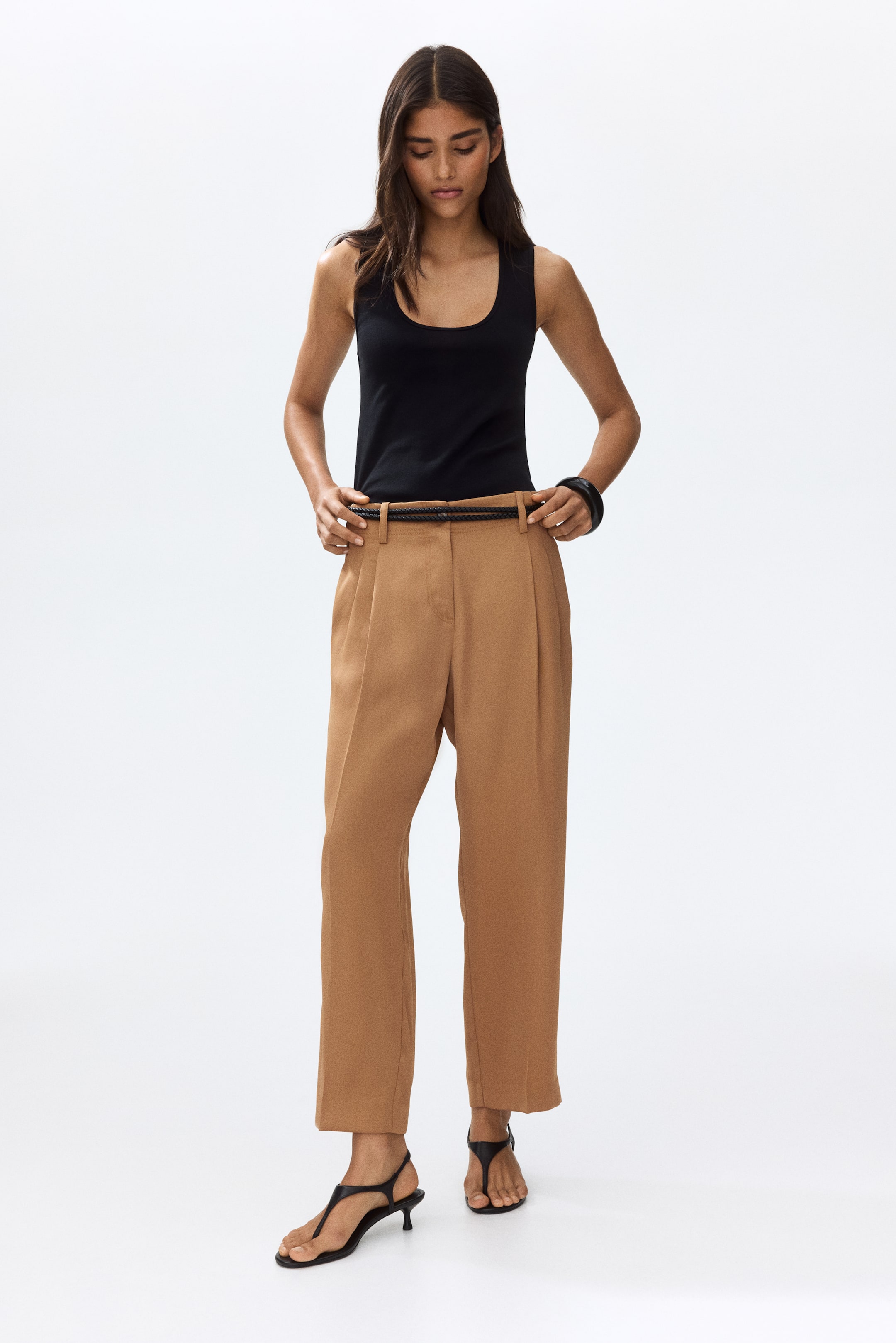 View larger image: "The model is wearing relaxed-fit tan trousers with a gentle drape, barrel legs, front pleats, and noticeable front creases. These trousers come with diagonal side pockets, and the model is seen adjusting the waistband, showcasing a black braided belt."