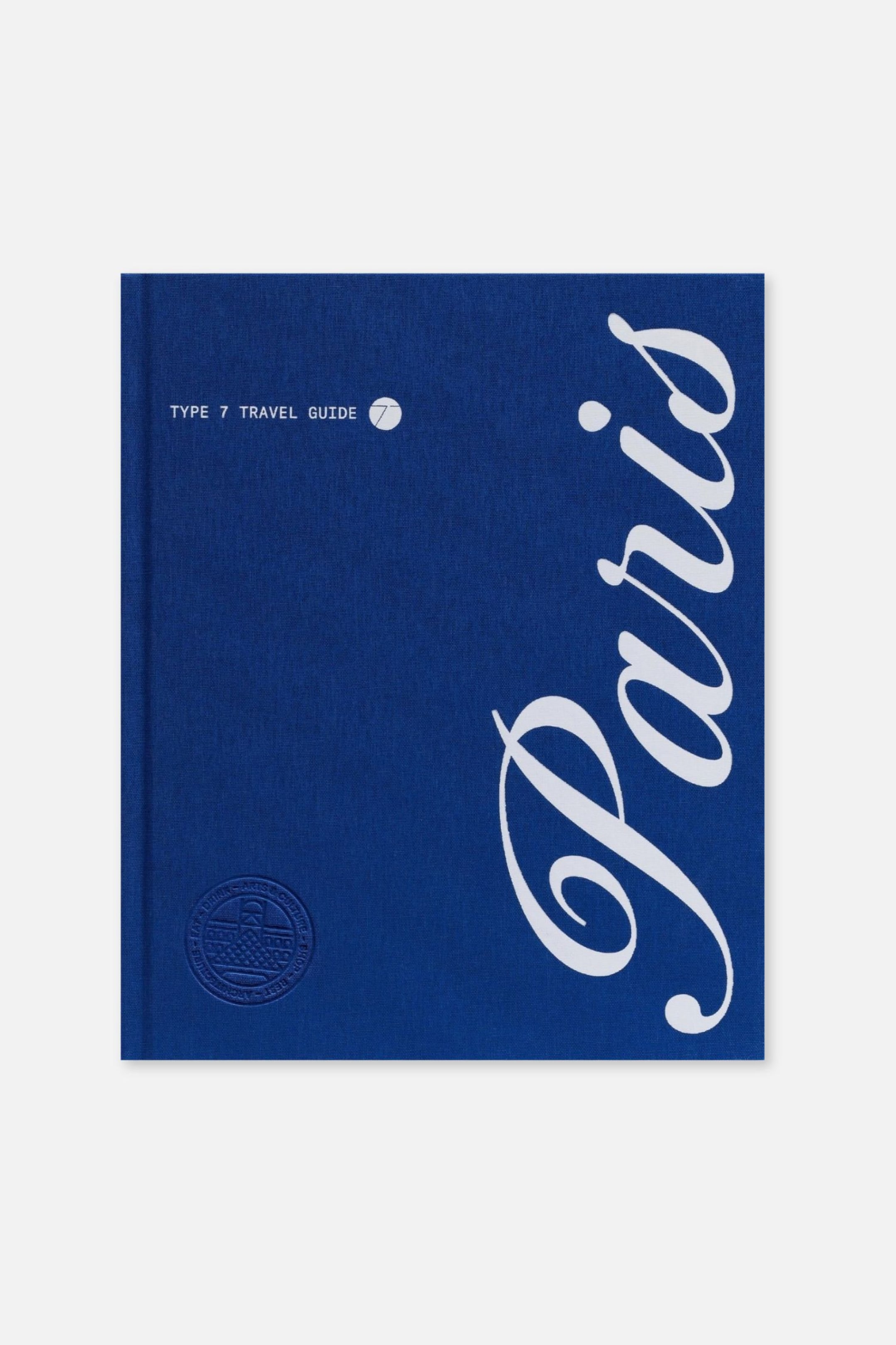 Agrandir l'image: Un livre relié en tissu bleu est présenté de face, avec le mot « Paris » écrit en grand en lettres verticales blanches sur la droite. La couverture affiche le texte « TYPE 7 TRAVEL GUIDE » à côté d’un petit logo circulaire en haut à gauche, avec un sceau circulaire embossé dans le coin inférieur gauche.