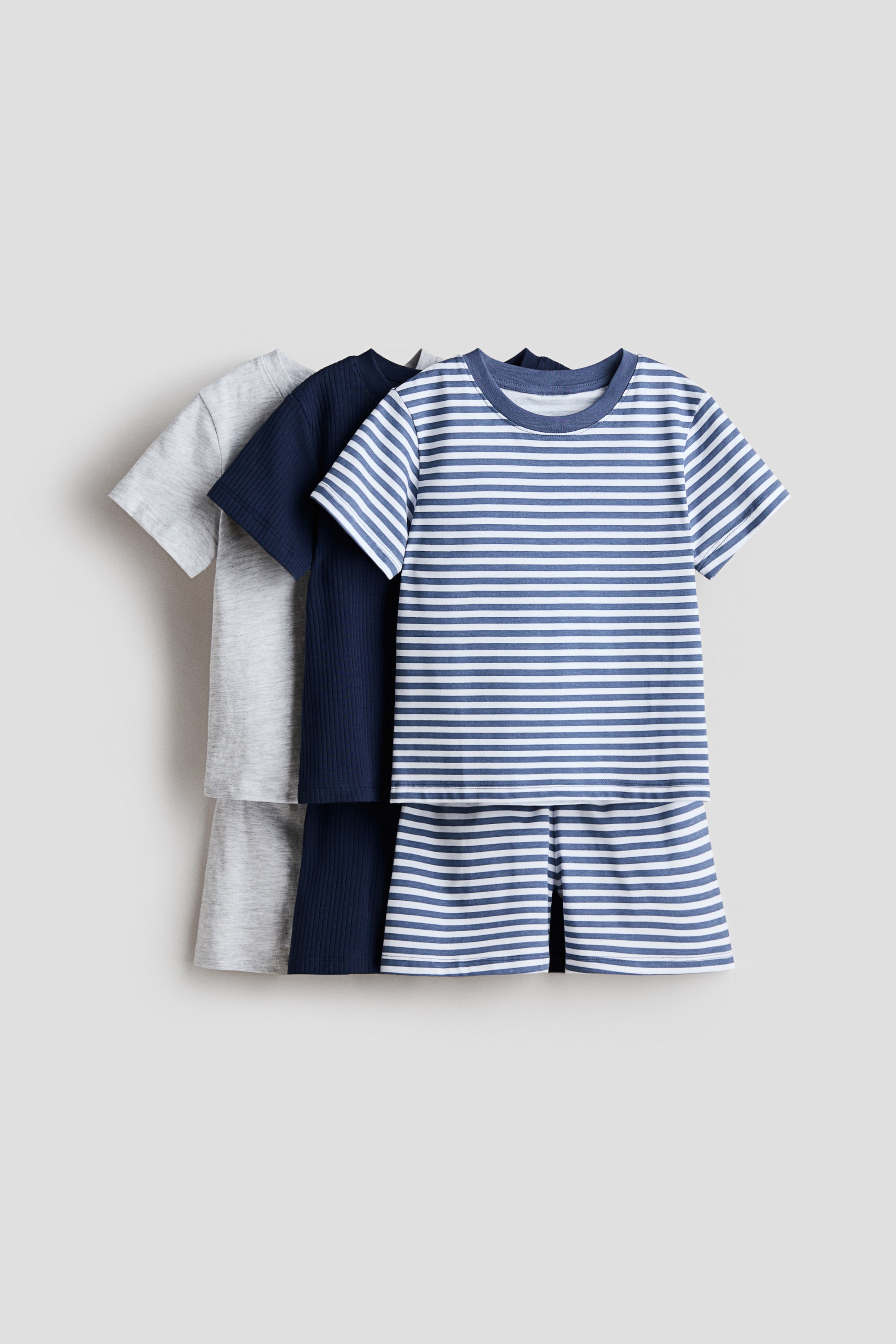이미지 확대 보기: 파자마 세트 세 개가 쌓여 있으며, 각 세트는 골지 넥라인이 있는 티셔츠와 신축성 있는 허리밴드가 있는 반바지로 구성되어 있습니다. 앞쪽 세트는 파란색과 흰색 줄무늬이며, 그 뒤로 네이비 블루 골지 세트와 연회색 세트가 보입니다.