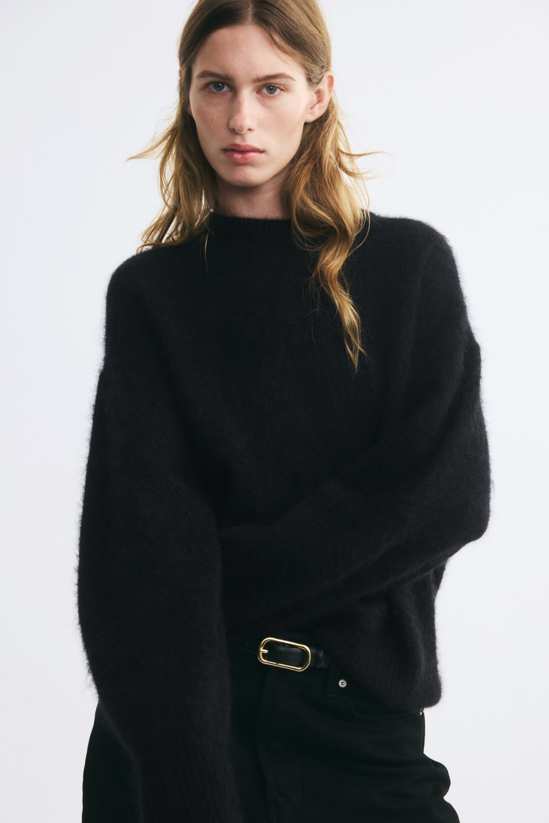 Ampliar la imagen: La modelo lleva puesto un suéter negro de punto, holgado y afelpado, con cuello redondo, hombros caídos y mangas largas y amplias. El suéter está parcialmente metido a la cintura dentro de unos jeans negros sujetos con un cinturón con hebilla dorada.