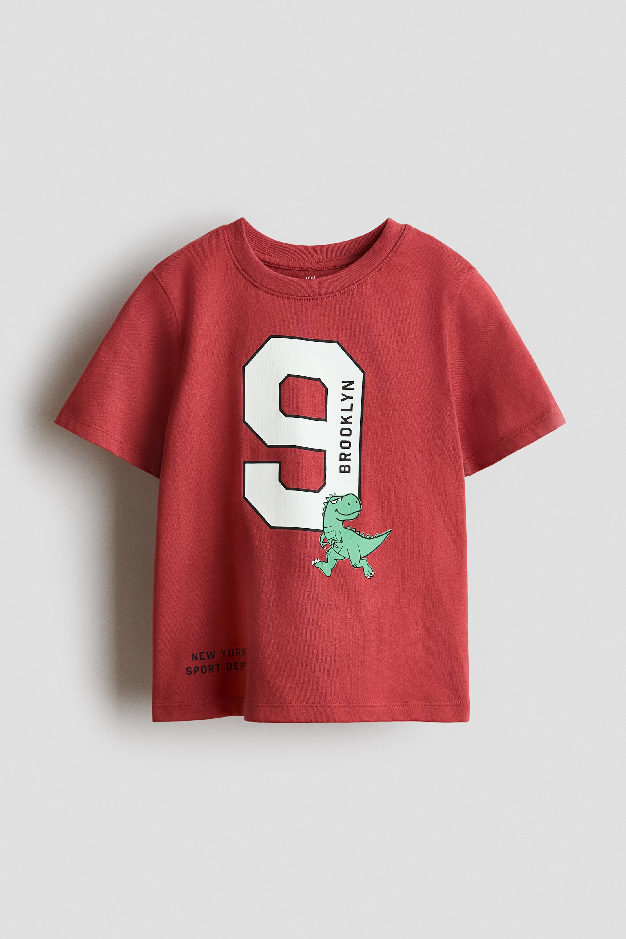 View larger image: A red short-sleeve t-shirt is showcasing its front, featuring a large white number nine graphic with a black outline, with 'BROOKLYN' printed vertically along its right side. A green cartoon dinosaur appears to be running from behind the bottom right of the nine, and small text reading 'NEW YORK SPORT DEP' is printed on the lower left front.