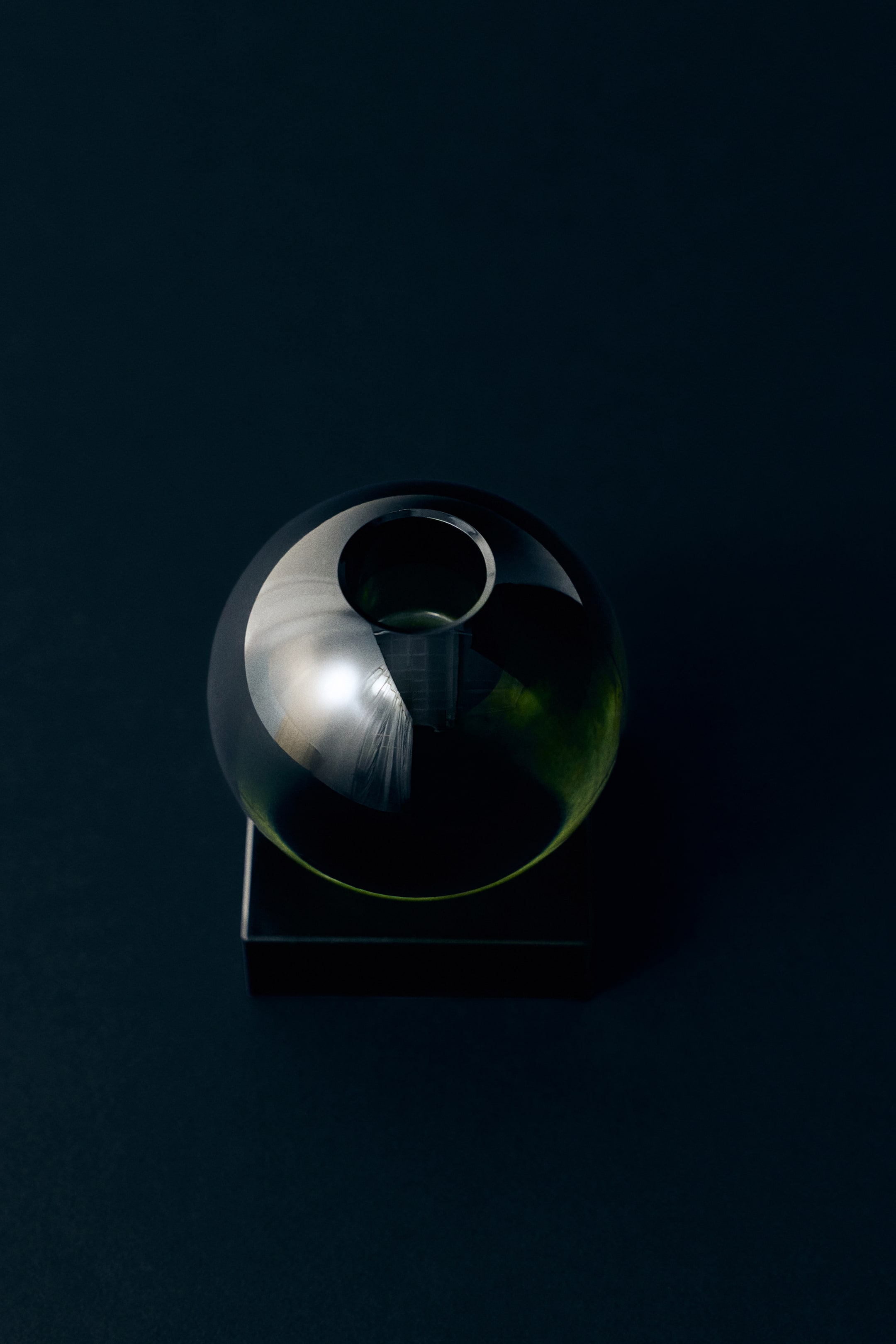 Visualizza l'immagine ingrandita: Un piccolo candelabro in vetro trasparente mette in risalto un design geometrico, caratterizzato da una sommità sferica con un'apertura circolare per una candela, poggiata su una base quadrata solida. Il prodotto è presentato da una vista frontale dall'alto, evidenziandone le superfici lisce e riflettenti.