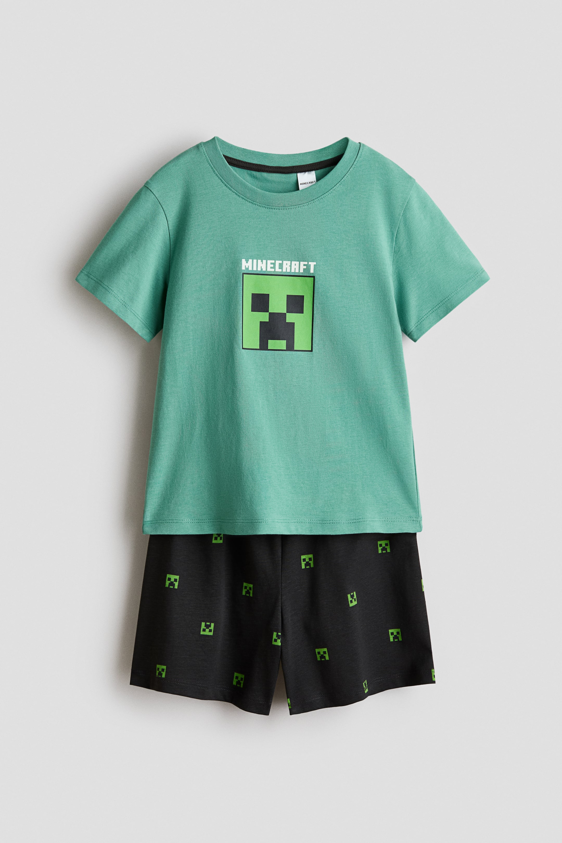 הצגת תמונה גדולה יותר: סט פיג'מה דו-חלקי של מיינקראפט מוצג מהחזית, הכולל חולצת טי ירוקה עם שרוולים קצרים ומכנסיים קצרים שחורים. החולצה מציגה לוגו "MINECRAFT" וגרפיקה גדולה של פנים מרובעים מפוקסלים בירוק, בעוד שהמכנסיים מציגים דפוס חוזר של פנים מרובעים מפוקסלים קטנים יותר בירוק.