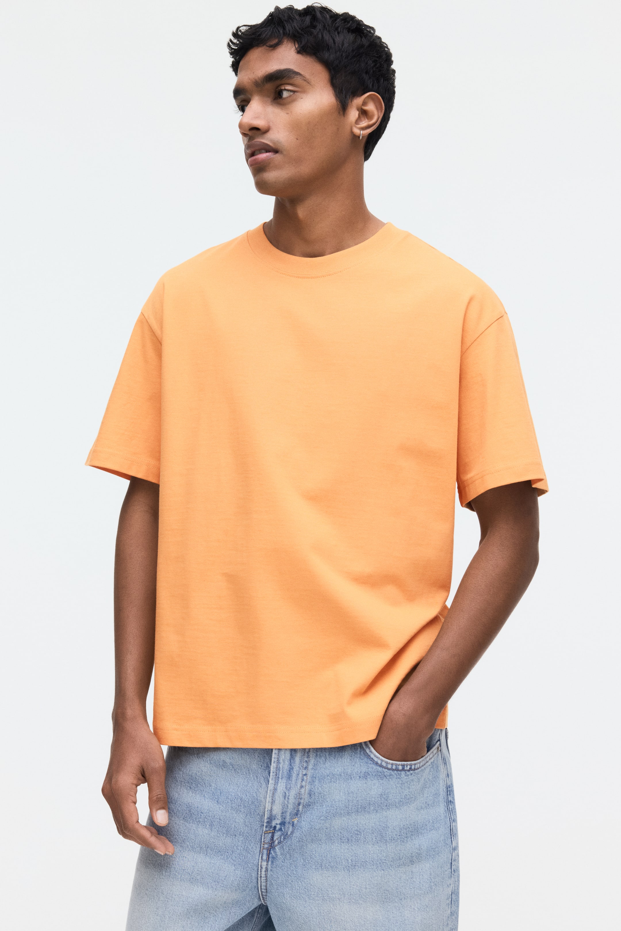 View larger image: The model is wearing an orange T-shirt featuring a round, ribbed neckline, dropped shoulders, and a straight-cut hem. This loose-fit T-shirt is styled with light blue jeans, with one of the model's hands casually placed in a front jean pocket.