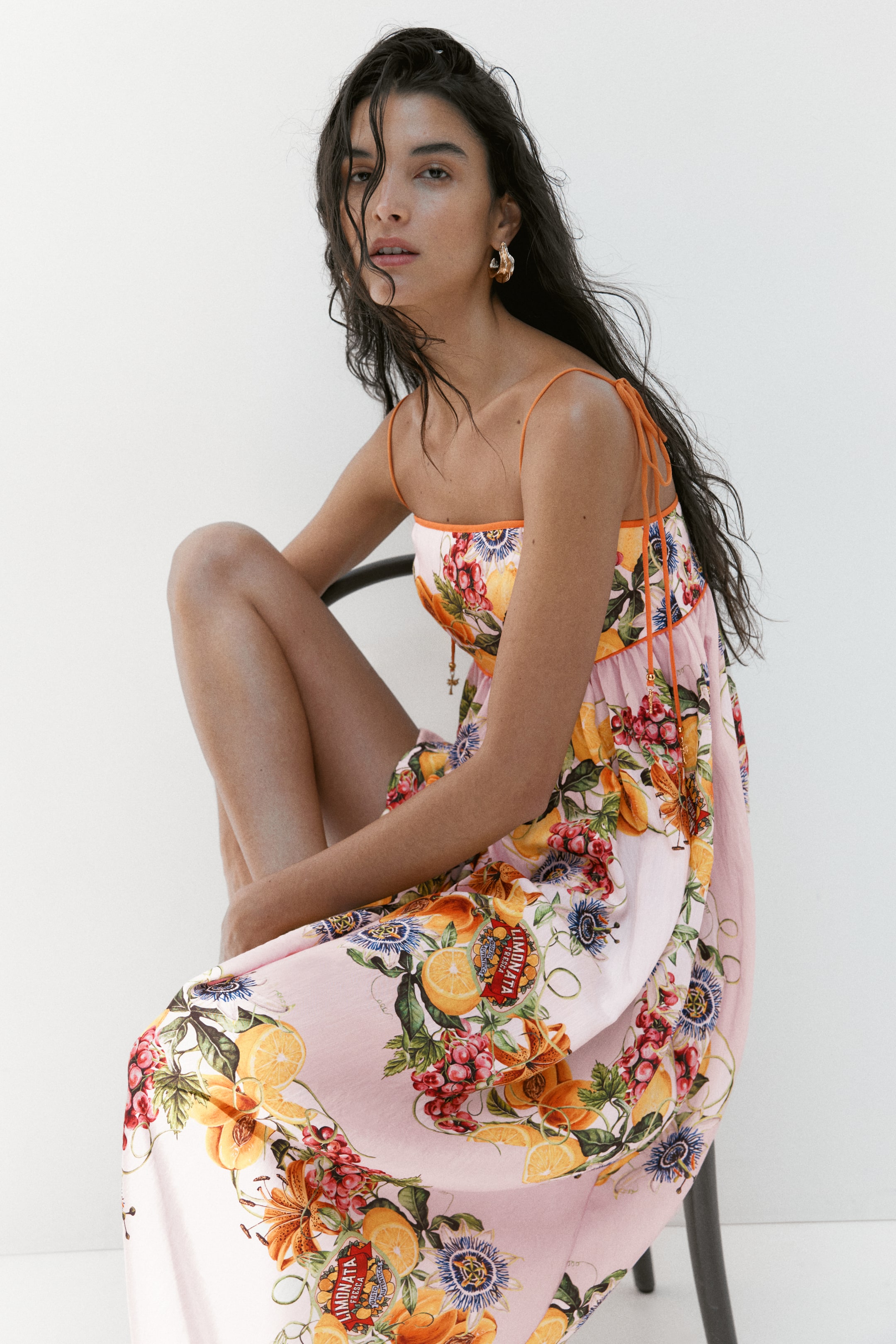 View larger image: The maxi dress showcases a soft pink fabric adorned with vibrant oranges, grapes, passion flowers, and "Limonata Fresca" labels. It features delicate orange spaghetti straps, a square neckline, and an empire waistline accentuated by orange ties with small gold bead accents, all visible from a front-side perspective.