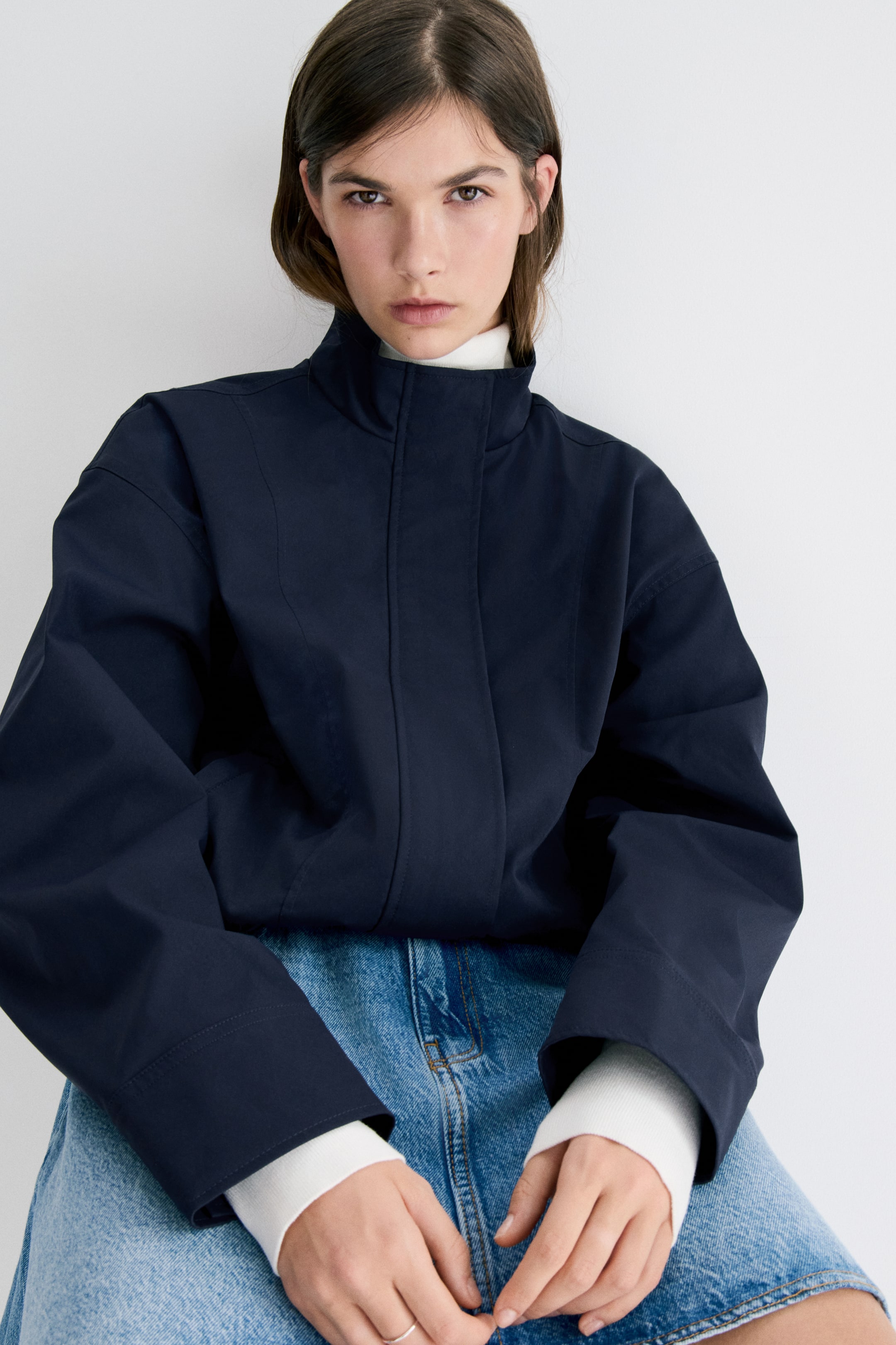 Xem ảnh lớn hơn: Một chiếc áo khoác màu xanh đậm đang được mặc, có cổ cao đứng và hàng khuy trước được giấu kín. Tay áo rộng kéo dài qua cổ tay có gân màu trắng, và các chi tiết đường may dọc định hình phía trước.