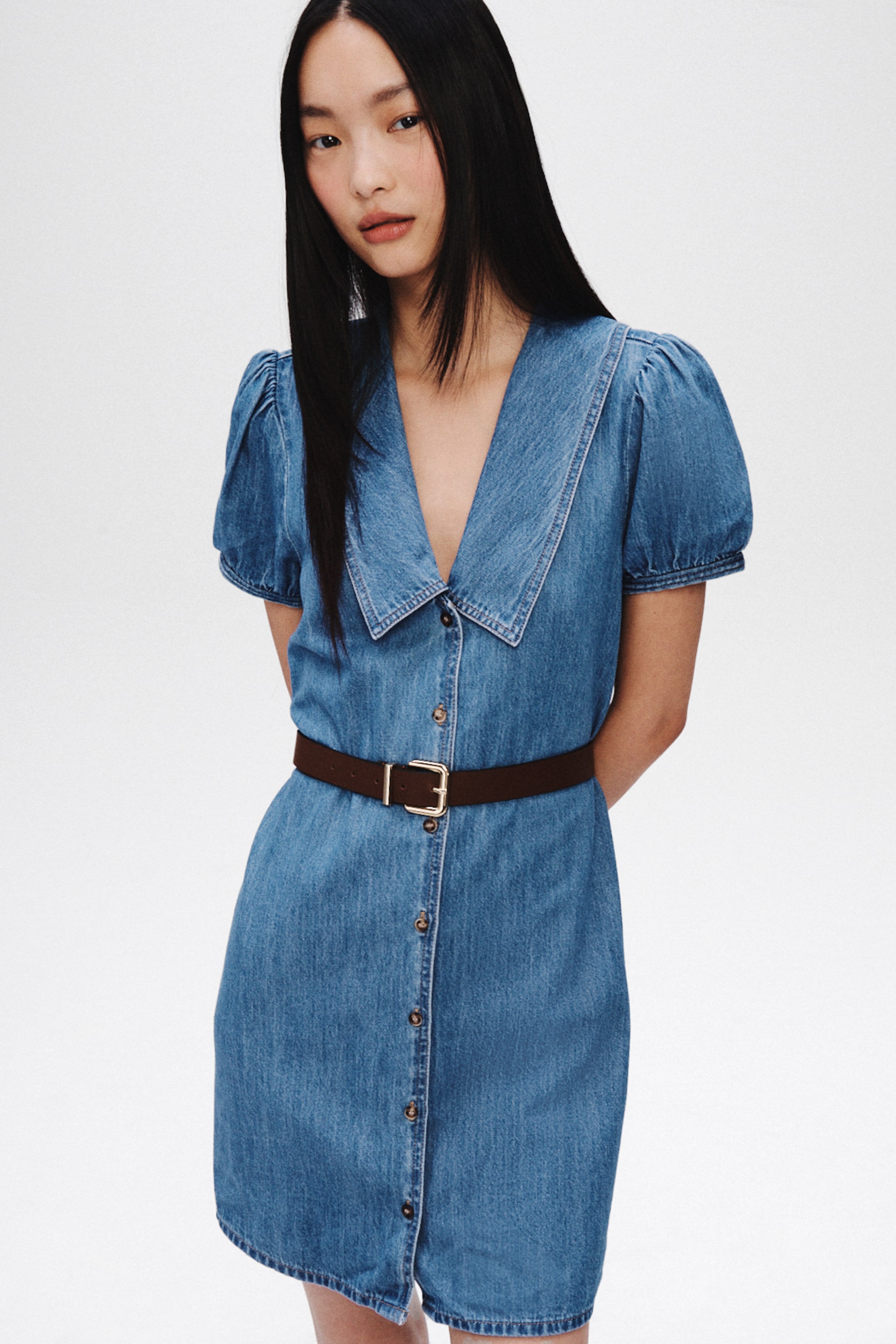 Xem ảnh lớn hơn: Chiếc váy mini denim màu xanh nhạt có cổ rộng nhọn, tay ngắn phồng và phần trước cài khuy toàn bộ với các nút màu nâu nhạt. Một chiếc thắt lưng màu nâu đậm với khóa mạ vàng siết eo váy.