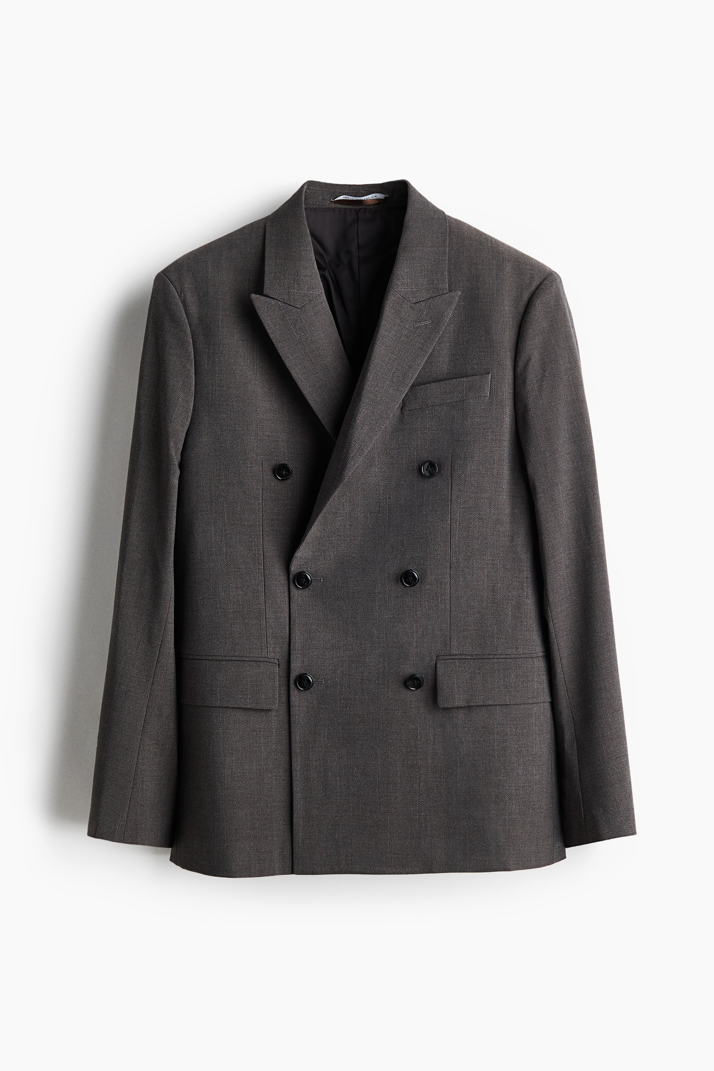 Una chaqueta gris de doble botonadura con solapas de pico, un bolsillo en el pecho y bolsillos delanteros con solapa, llevada por la modelo sobre un jersey oscuro en capas y pantalones grises a juego. La chaqueta presenta una silueta relajada, y la modelo aparece de pie con las manos en los bolsillos del pantalÃ³n.