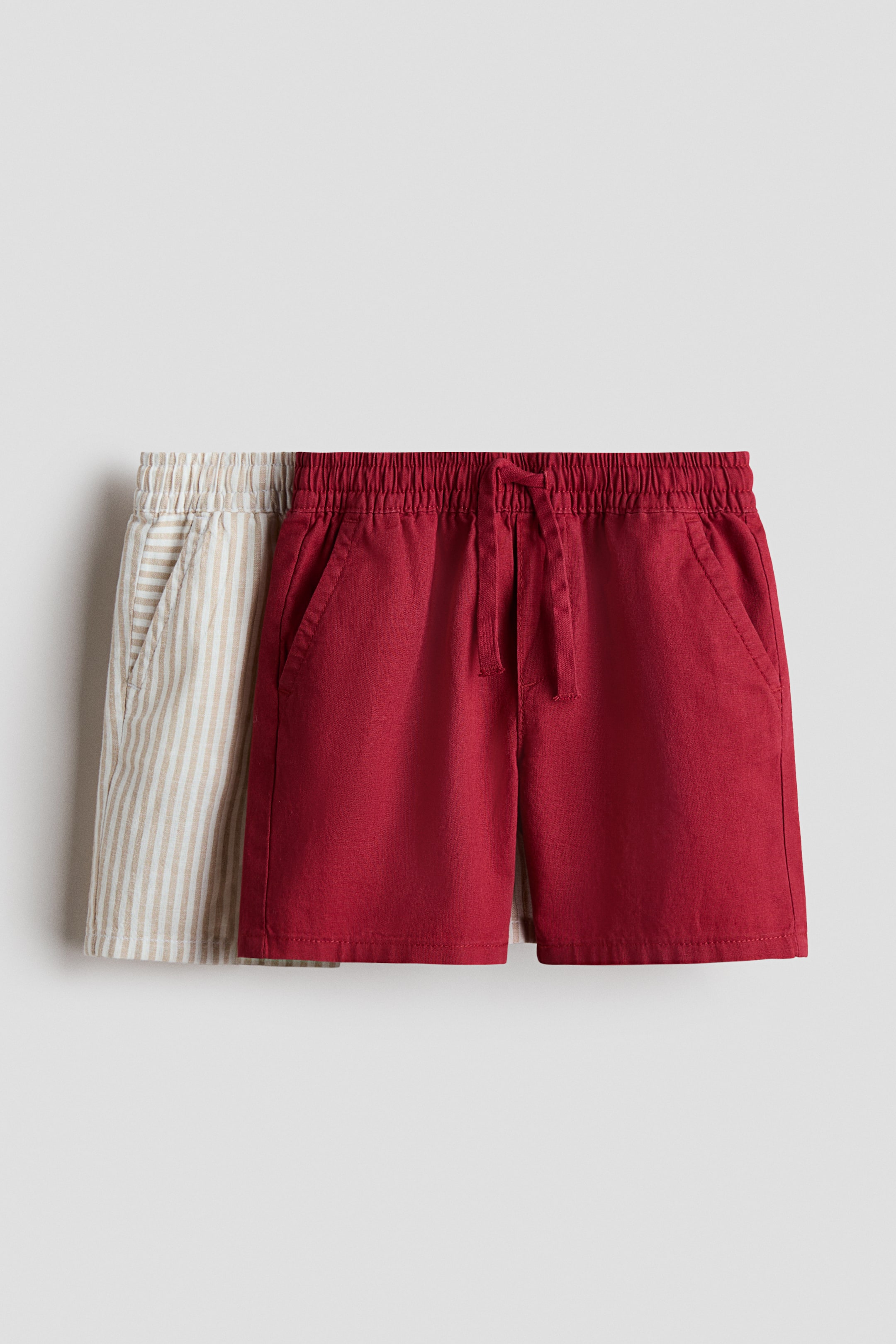 Agrandir l'image: Deux paires de shorts sont posées à plat, mettant en valeur leur devant. La paire à l'avant présente un tissu rouge foncé uni, avec une taille élastiquée munie d'un cordon de serrage et des poches latérales inclinées. La deuxième paire, partiellement visible à l'arrière, montre un tissu beige à rayures verticales, présentant également une taille élastiquée et des poches latérales inclinées.