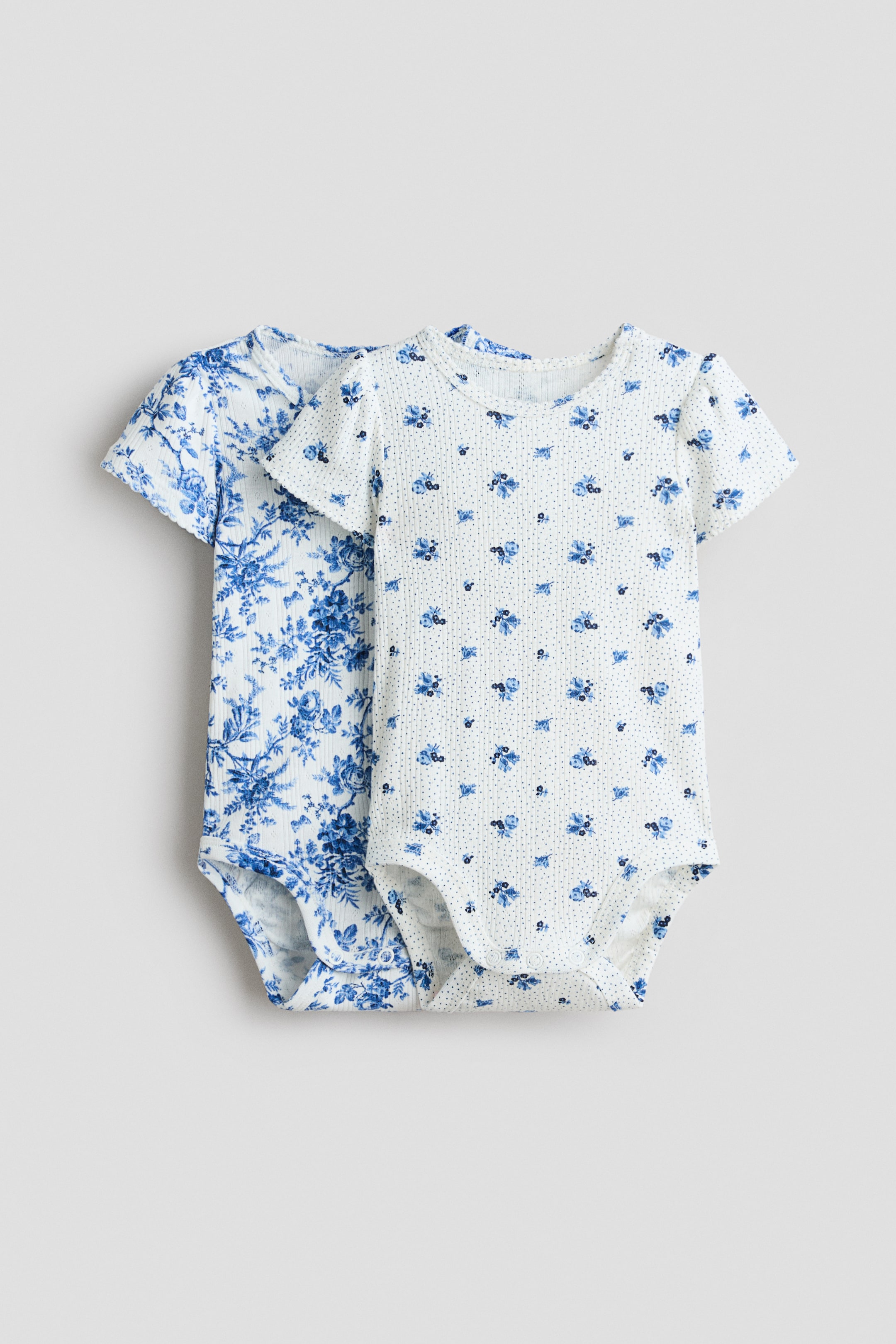 Grösseres Bild ansehen: Zwei Baby-Bodys werden präsentiert, mit der Vorderseite nach vorne. Der vordere Body besteht aus weißem Rippstoff und zeigt kleine blaue Blumenmotive und winzige blaue Punkte, kurze Ärmel und einen runden Halsausschnitt. Der zweite Body, leicht dahinter positioniert, besteht ebenfalls aus weißem Rippstoff und zeigt einen dichten blauen Blumenprint sowie kurze Ärmel.