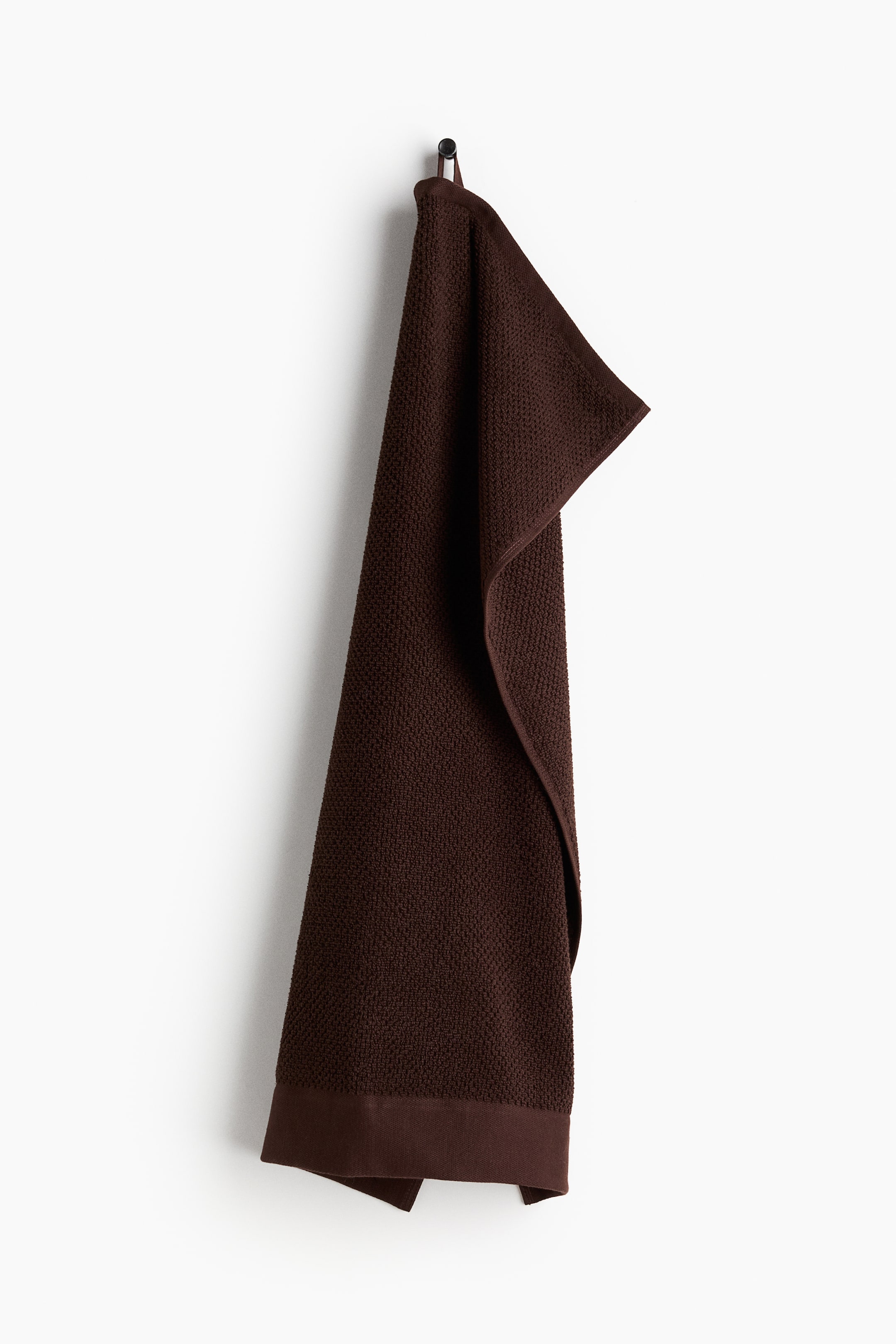 View larger image: A dark brown hand towel is hanging vertically, showcasing its textured surface. It is suspended by a fabric loop at its top end, and a smooth band finishes the bottom edge.
