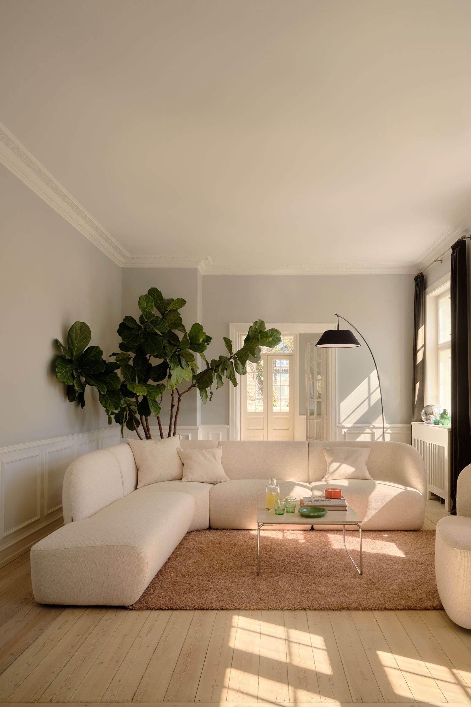 Een modulaire hoeksofa bekleed met een gestructureerde crèmekleurige stof staat naar voren gericht en toont een laag profiel met afgeronde randen en naadloze, blokachtige kussens. De sofa is opgesteld in een L-vorm met een chaise longue die links naar voren uitsteekt onder een lage, geïntegreerde rugleuning.