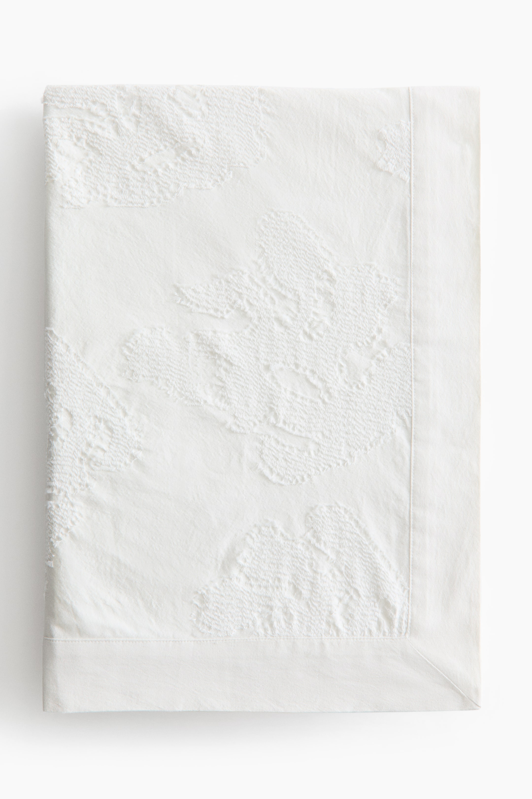 拡大画像を表示: 白い折りたたまれた織物が平らに広げられており、主な表面には抽象的な有機形状のテクスチャーのある隆起したパターンが描かれています。右端と下端には滑らかで無地の縁が見えます。