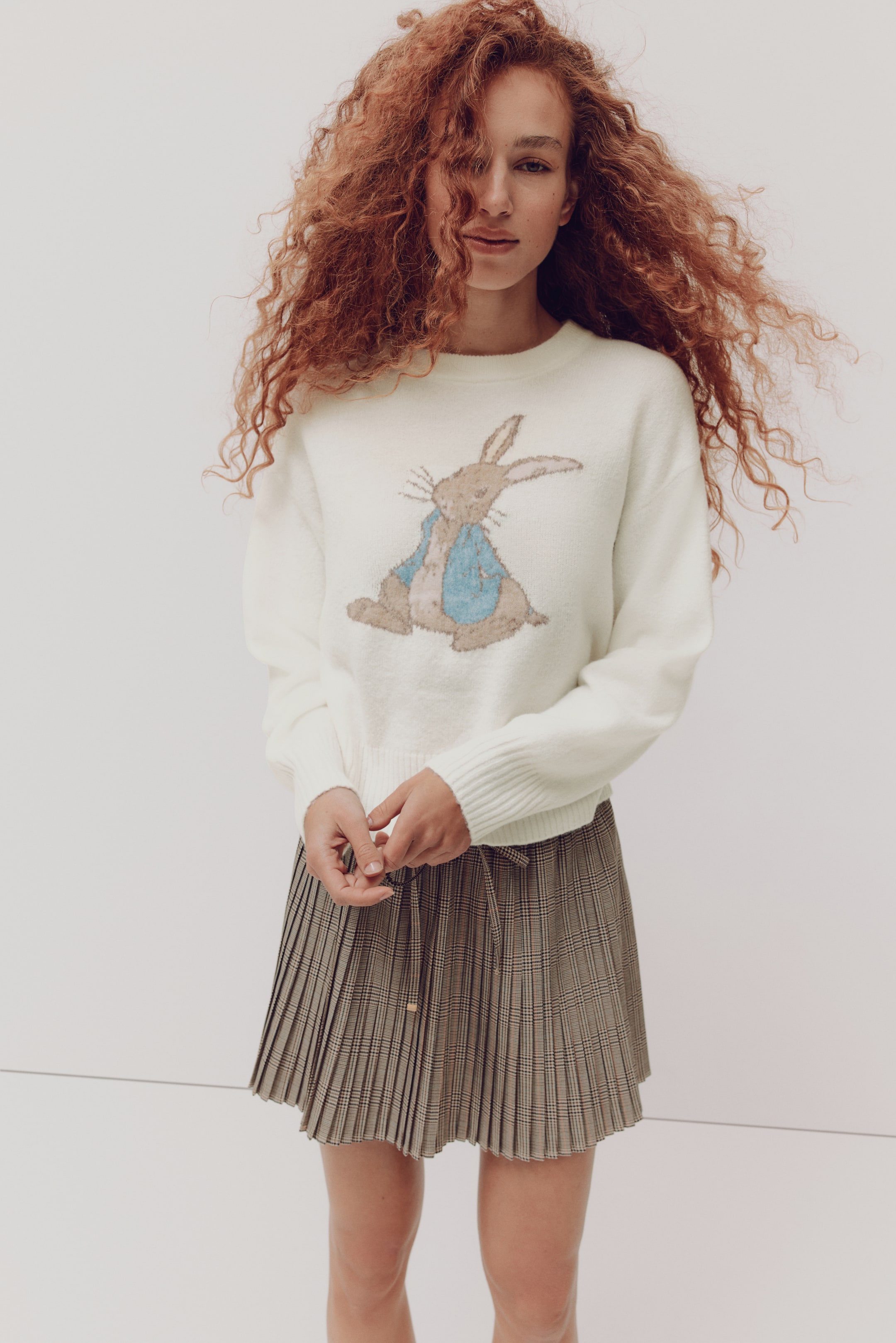 拡大画像を表示: オフホワイトのクルーネックセーターに、ブラウンとブルーのピーターラビットのイラストがあしらわれ、ウエストにドローストリングの結び目が見えるブラウン×ブラックのチェック柄プリーツミニスカートと合わせられています。