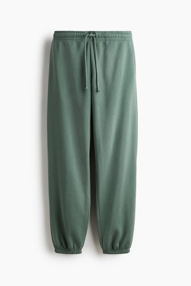 Un par de joggers verde apagado se presenta desde el FRENTE. Los joggers cuentan con una tela de sudadera, una cintura fruncida elástica con un cordón visible atado en el centro y elásticos cubiertos en los dobladillos. - 1