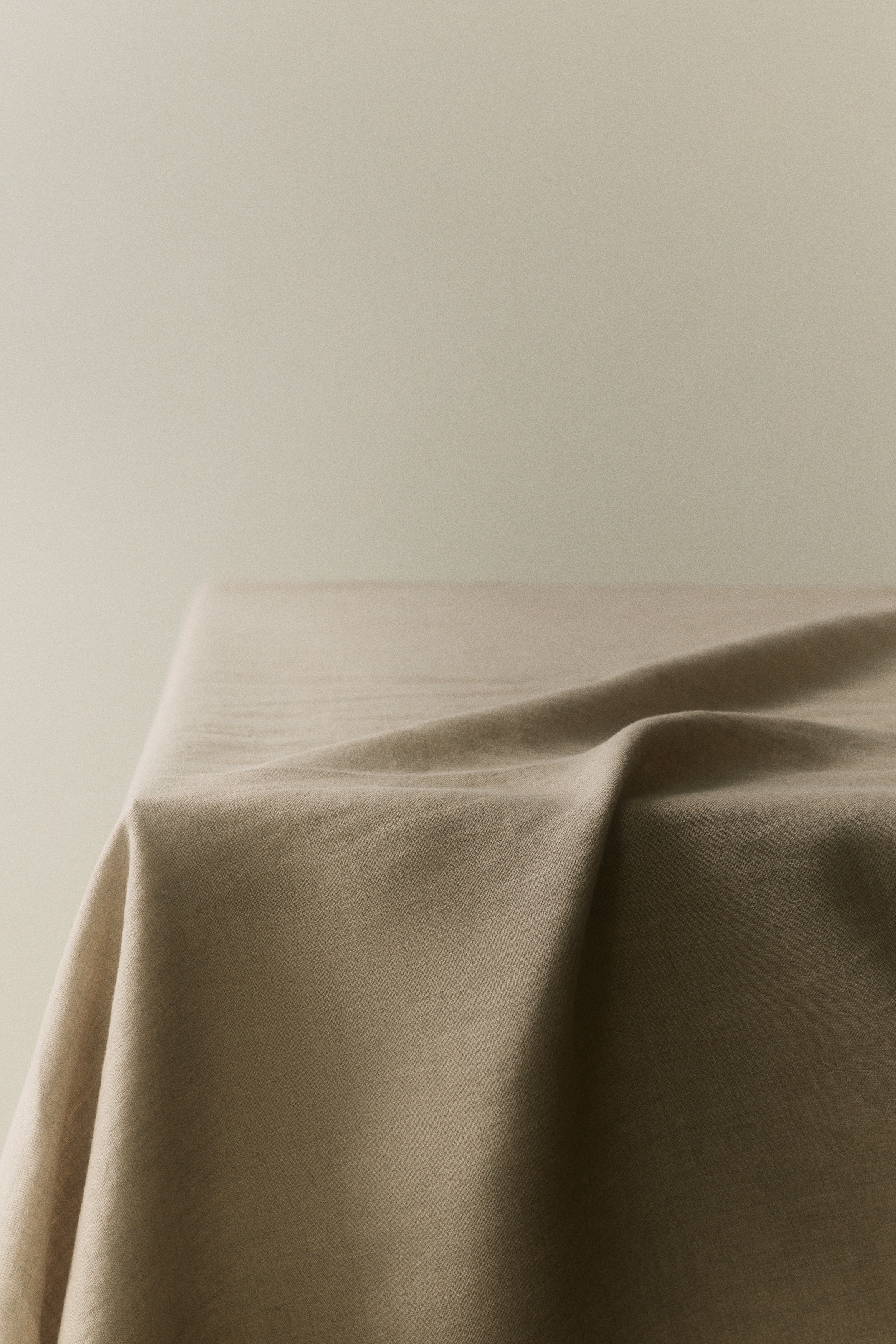 Een licht beige tafellaken in linnenweefsel ligt plat, gezien vanaf de voorkant. Het heeft een gevouwen en gestikte zoom langs de rechter- en onderrand.